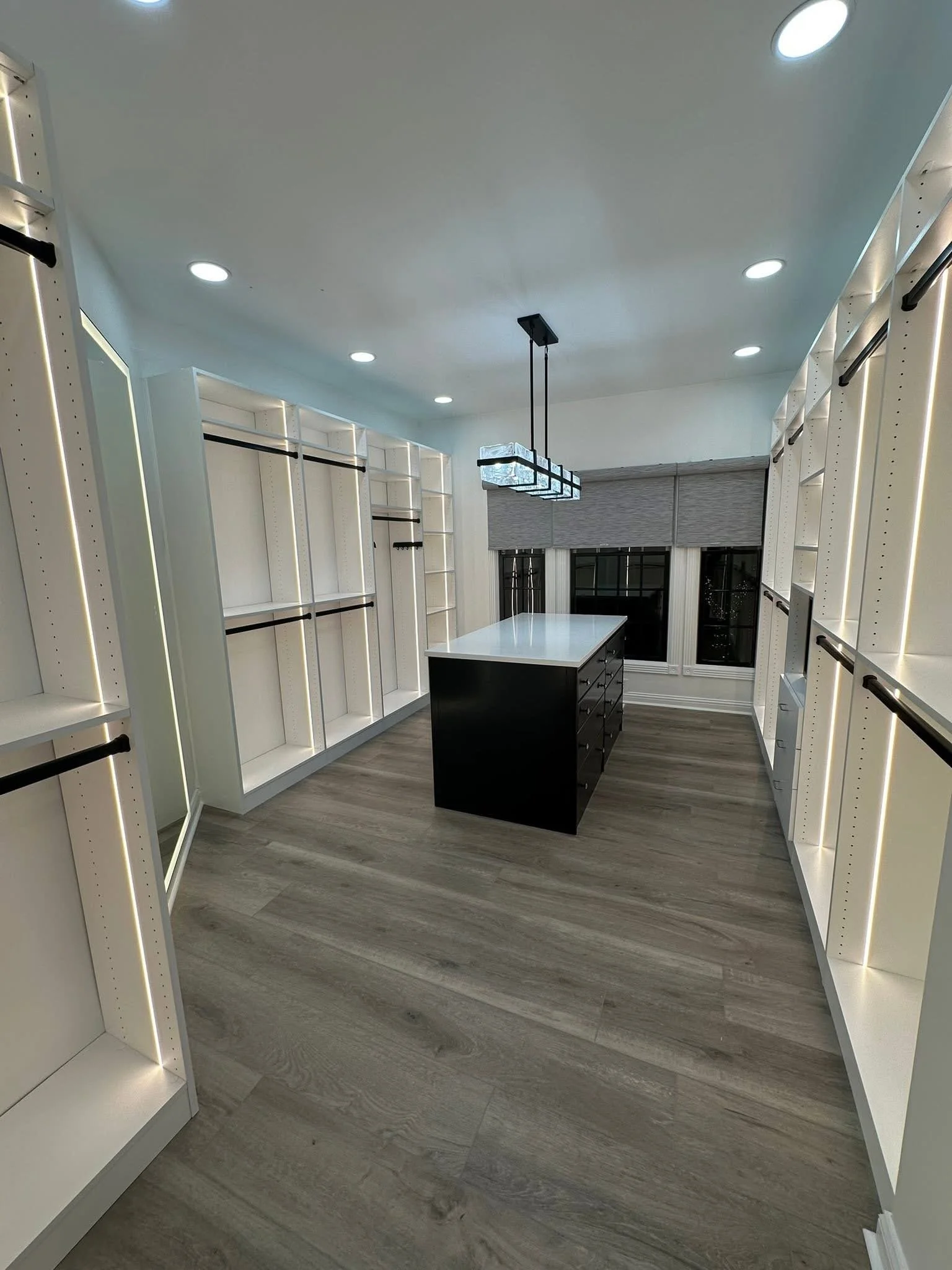 An empty walk-in closet with white shelving on both sides, a central black island with drawers, and a window with closed gray blinds. Recessed ceiling lights illuminate the room.
