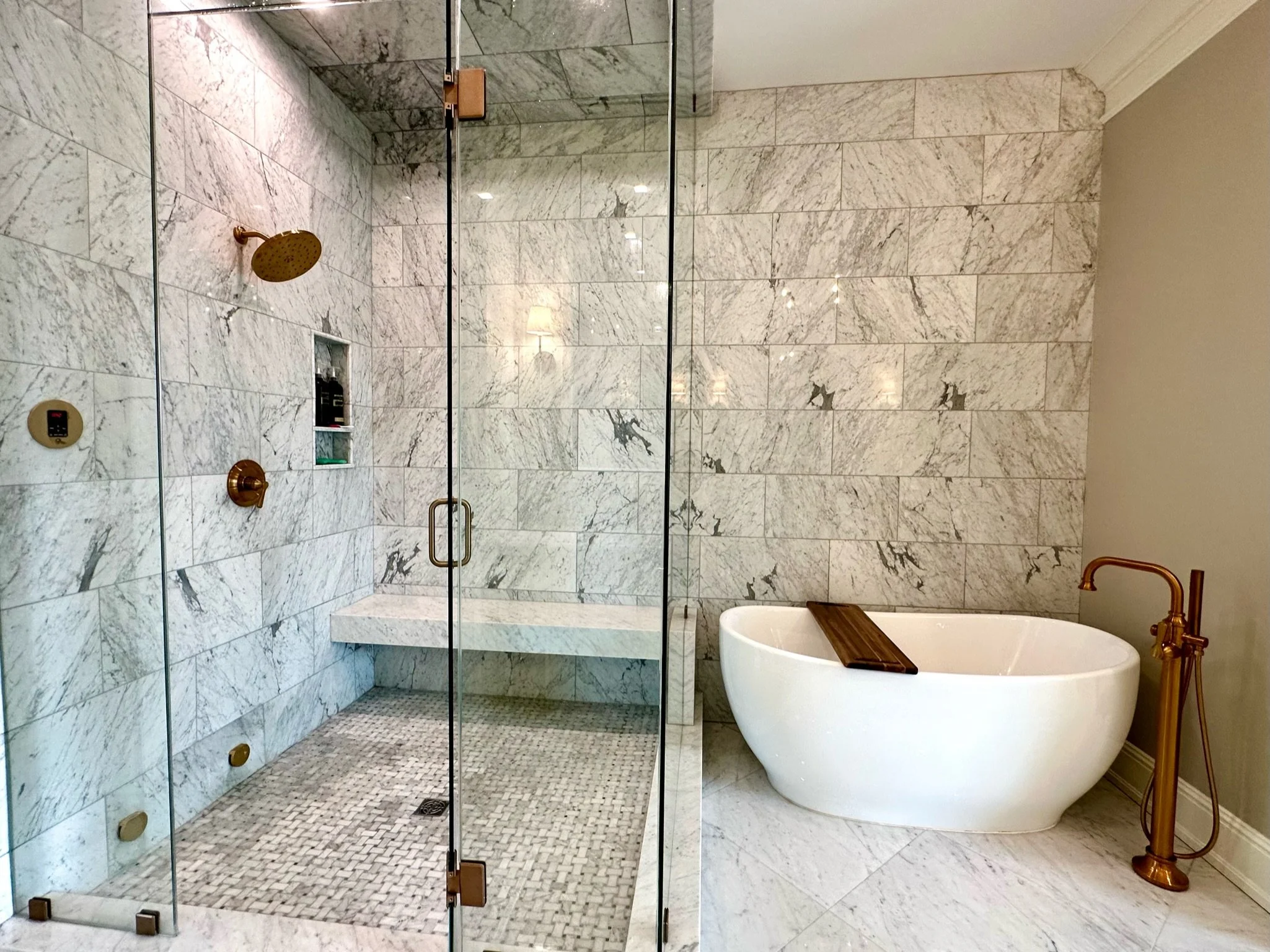 A spacious bathroom shower area with marble tile walls and floor, a glass door, a wall-mounted showerhead, a built-in niche for toiletries, and a separate freestanding bathtub near a rustic wooden basin with a gold-colored faucet and stand.