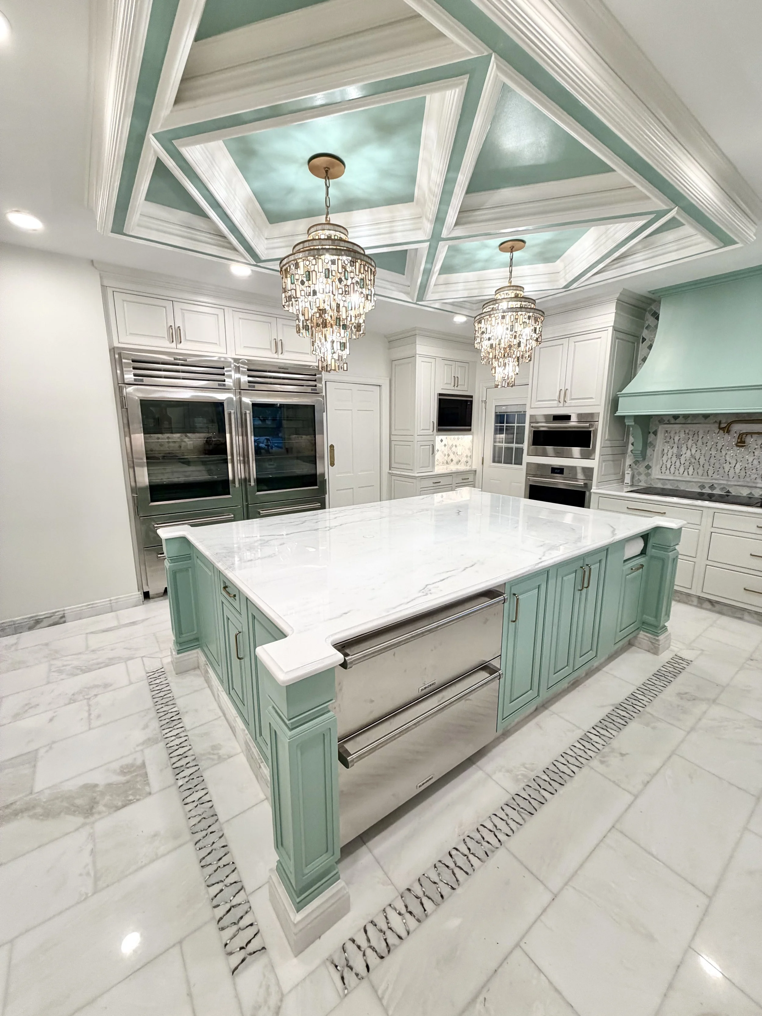 A kitchen with a large marble island with mint green cabinets, white marble flooring, and two ornate chandeliers hanging from a decoratively painted coffered ceiling.