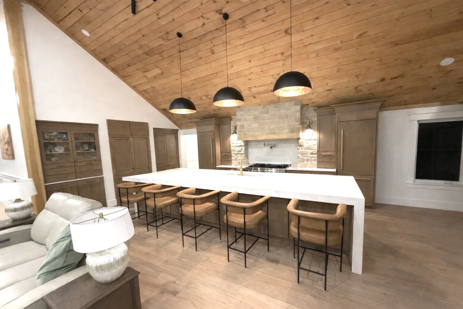 Modern kitchen with wooden ceiling, white island, black pendant lights, and brown cabinets.