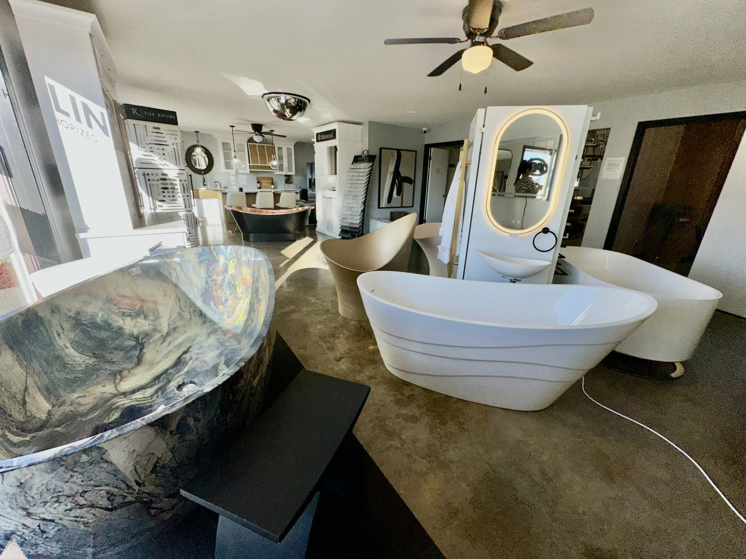 Interior view of a modern home decor store featuring a marble bathtub, white chairs, a large fancy mirror with LED lighting, ceiling fans, and various wall decorations.