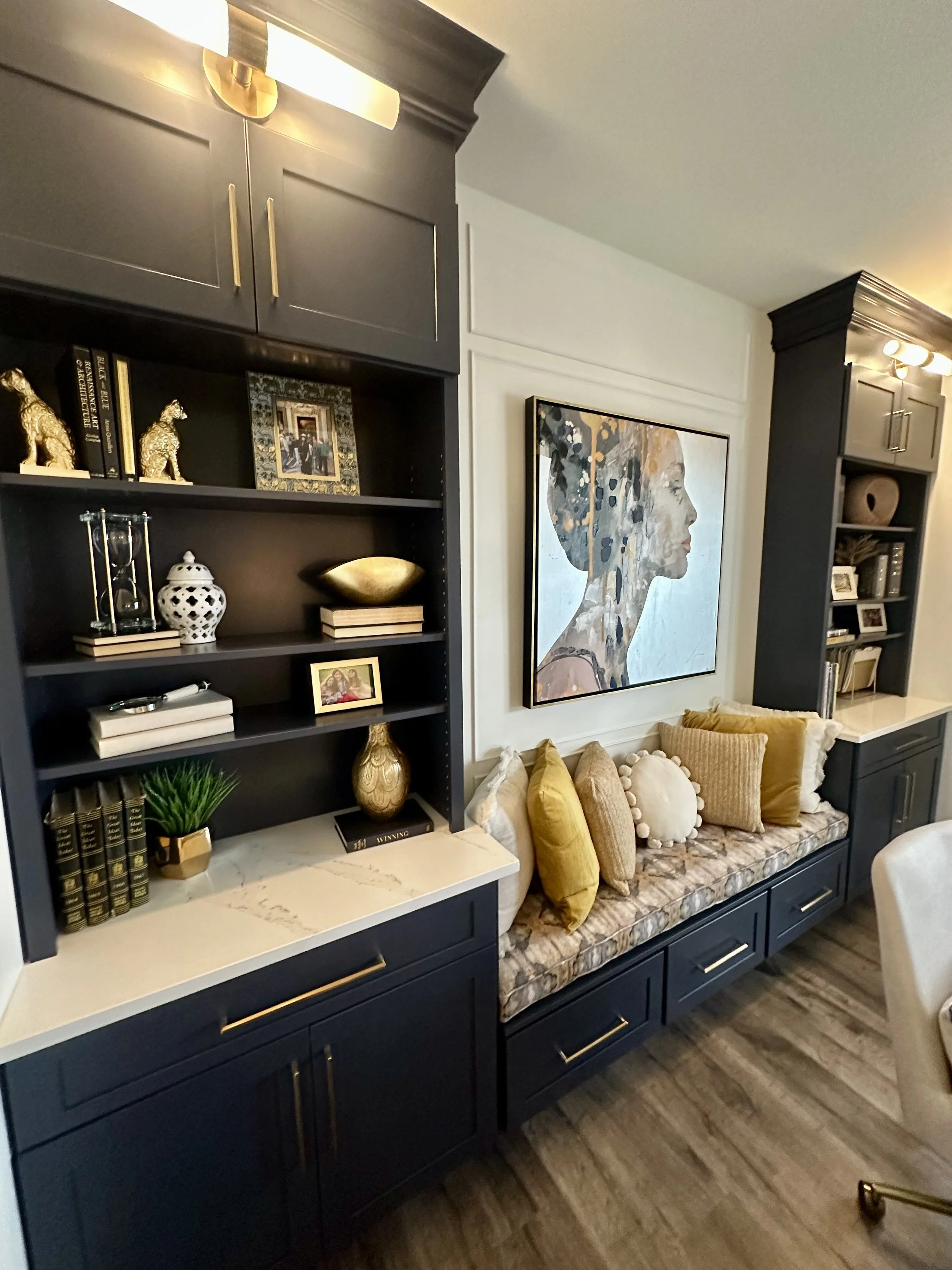 Living room with dark blue built-in shelves, gold hardware, and a marble-topped cabinet. Decorative items include gold horse figurines, books, framed photos, a gold bowl, and small plants. A large framed portrait of a woman hangs on the wall, and a cushioned bench with yellow and white pillows is beneath it, next to the shelving.