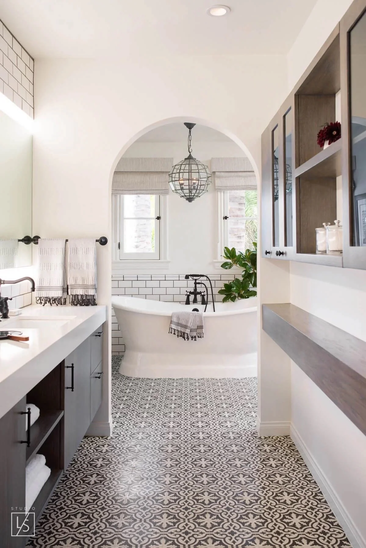 Bathroom with black and white patterned tile floor, white clawfoot bathtub near window, cabinets with black hardware, and decorative towels, window with roman shade, pendant light, and plants.