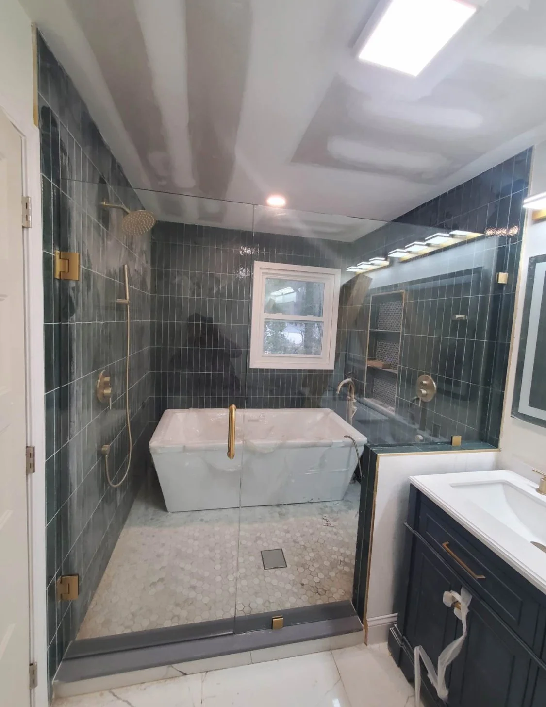 Modern bathroom with dark green tiled shower area, a white bathtub, and a window. The shower has gold fixtures, including a handrail, showerhead, and controls. The ceiling is partially finished with drywall patches, and there is a vanity with a white sink and dark cabinet.