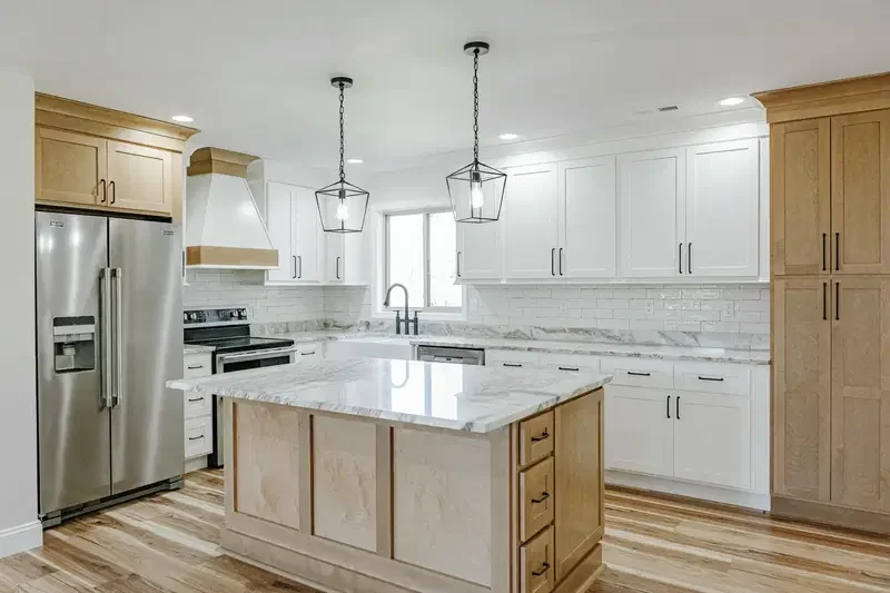 Modern kitchen with white cabinets, a center island with a marble top, stainless steel refrigerator, oven, and hood, wood flooring, and pendant lighting.