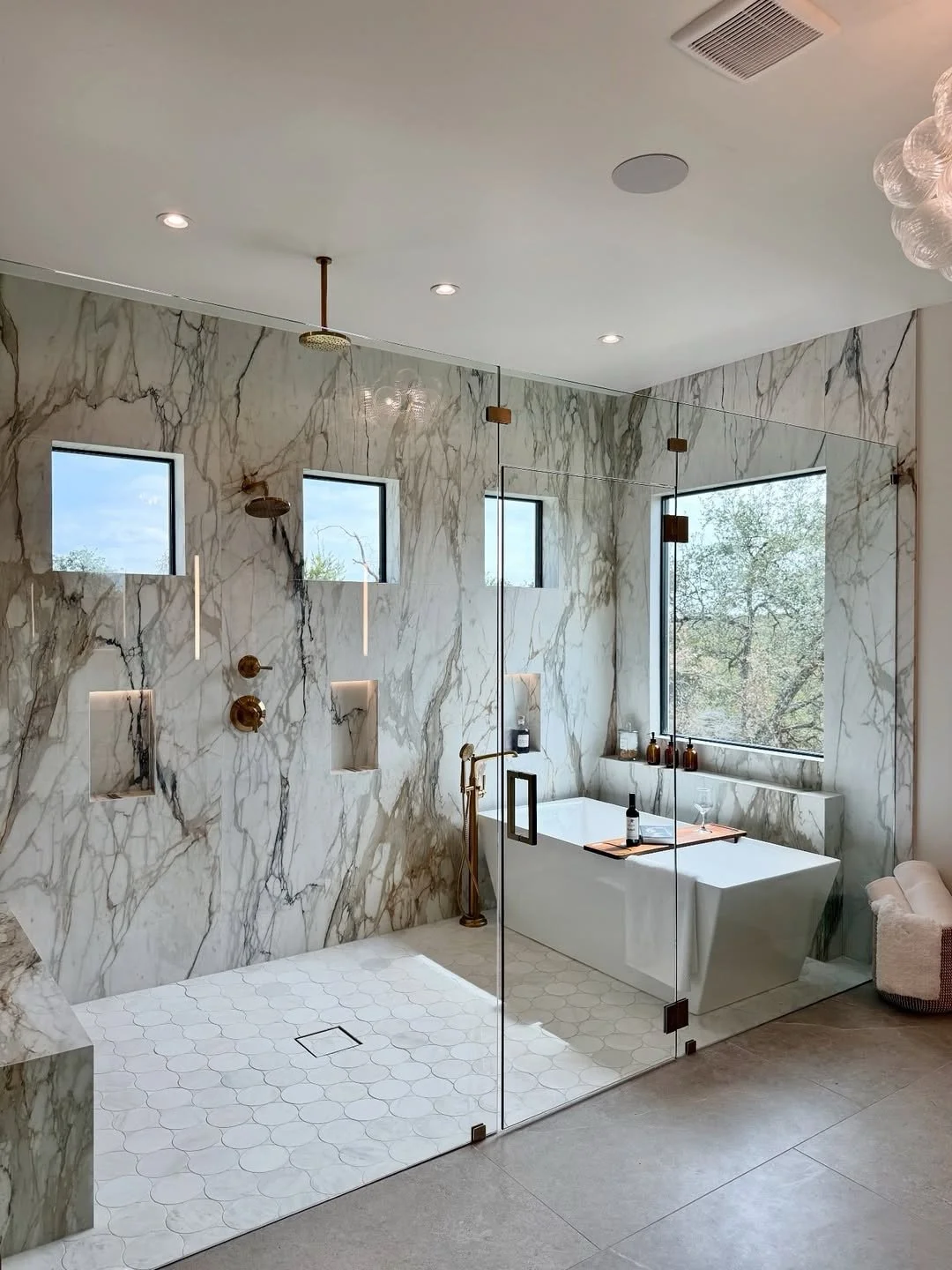 Modern bathroom with marble walls, walk-in shower with white hexagonal tiles, a freestanding bathtub with a large window, and minimal decor.