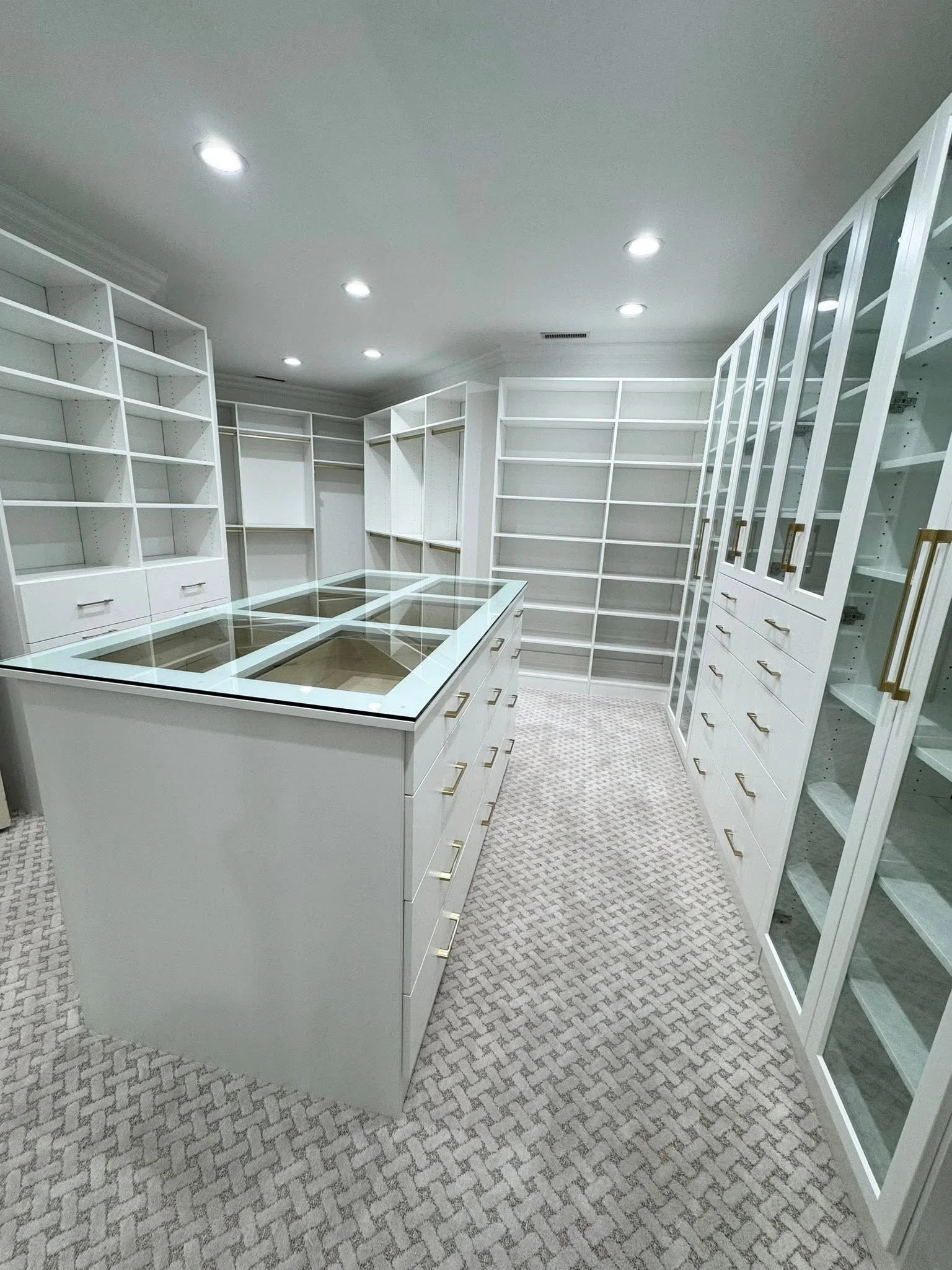 Empty white walk-in closet with glass-topped storage drawers, built-in shelves, and glass-front cabinets, carpeted floor, white walls, ceiling with recessed lighting.