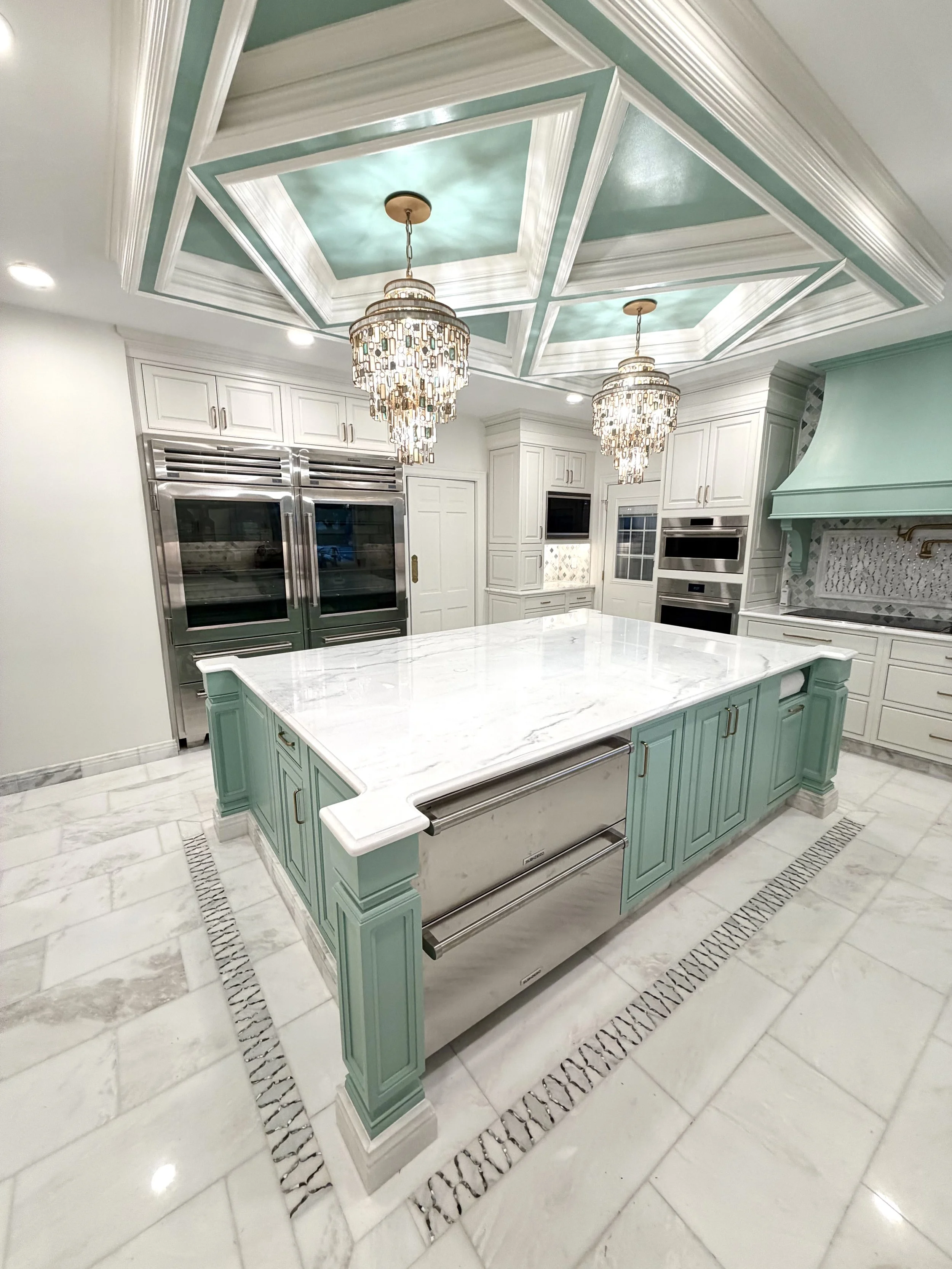 A luxurious kitchen with a pastel green kitchen island, white marble countertop, and elegant chandelier lighting fixtures hanging from a detailed coffered ceiling. The kitchen is equipped with modern stainless steel appliances, white cabinetry, and intricate tile flooring.