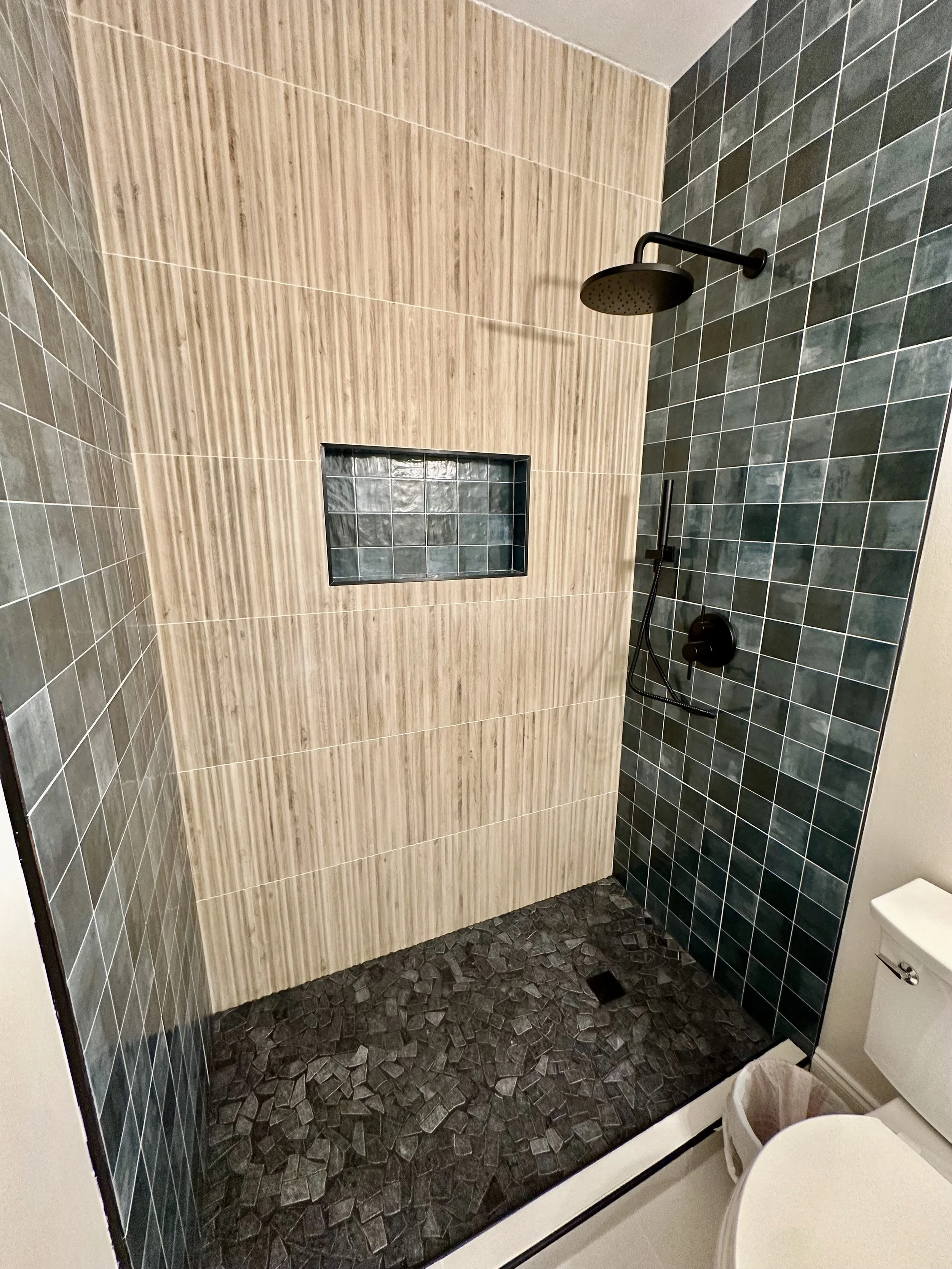 Modern walk-in shower with black rain shower head, black handheld shower, dark gray tiled walls, a niche with tiled backing, and a textured black tile floor.