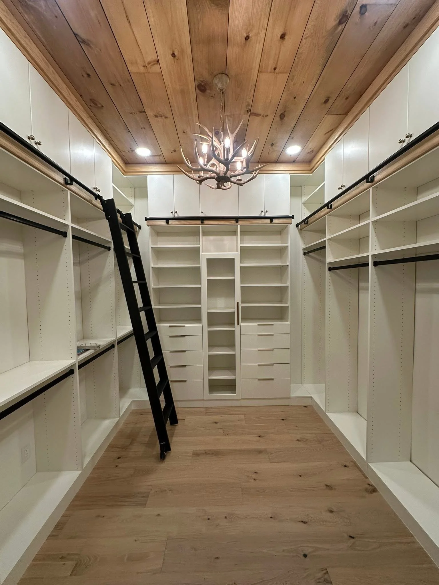 A walk-in closet with white bookshelves, black sliding closet door tracks, a black ladder, hardwood flooring, a wooden ceiling, and a chandelier with antler design.