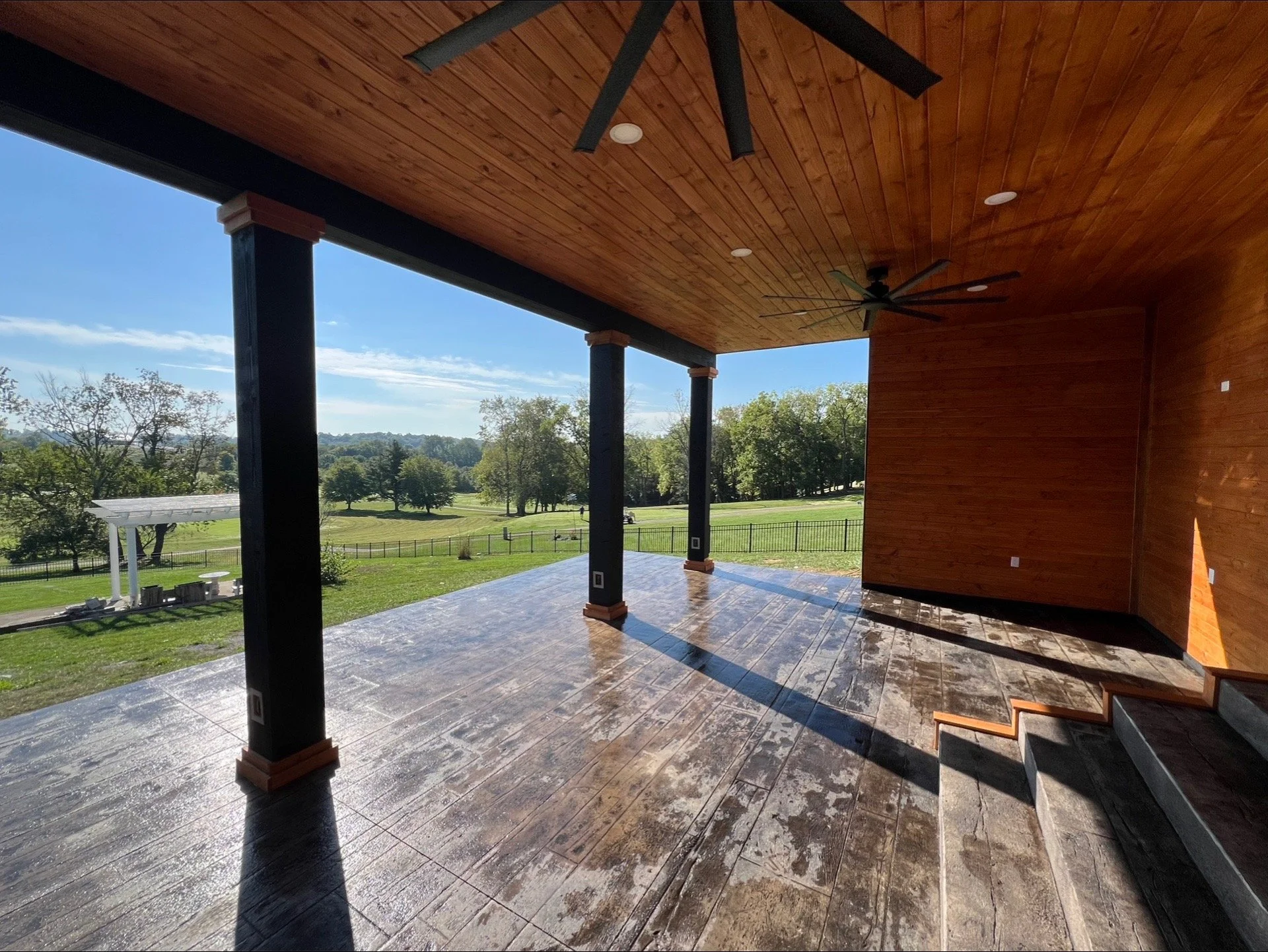 Covered patio area with wood-paneled ceiling, ceiling fans, and tiled floor, overlooking a lush green landscape with trees and a small white structure in the distance.