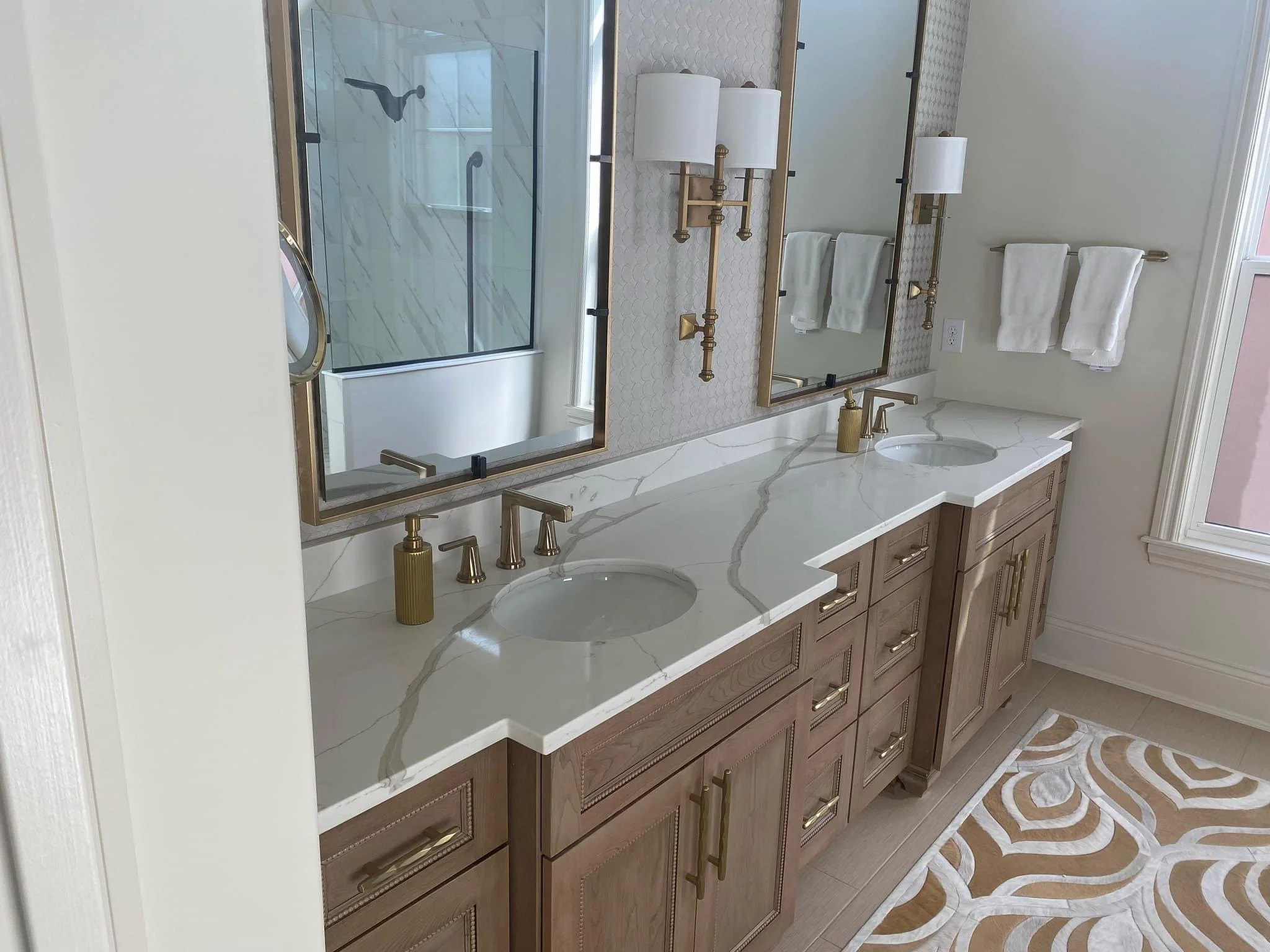 Bathroom vanity with double sinks, marble countertop, large mirror with gold framing, wall-mounted gold lighting fixtures, white towels on a towel rack, and a patterned area rug on wooden floor.