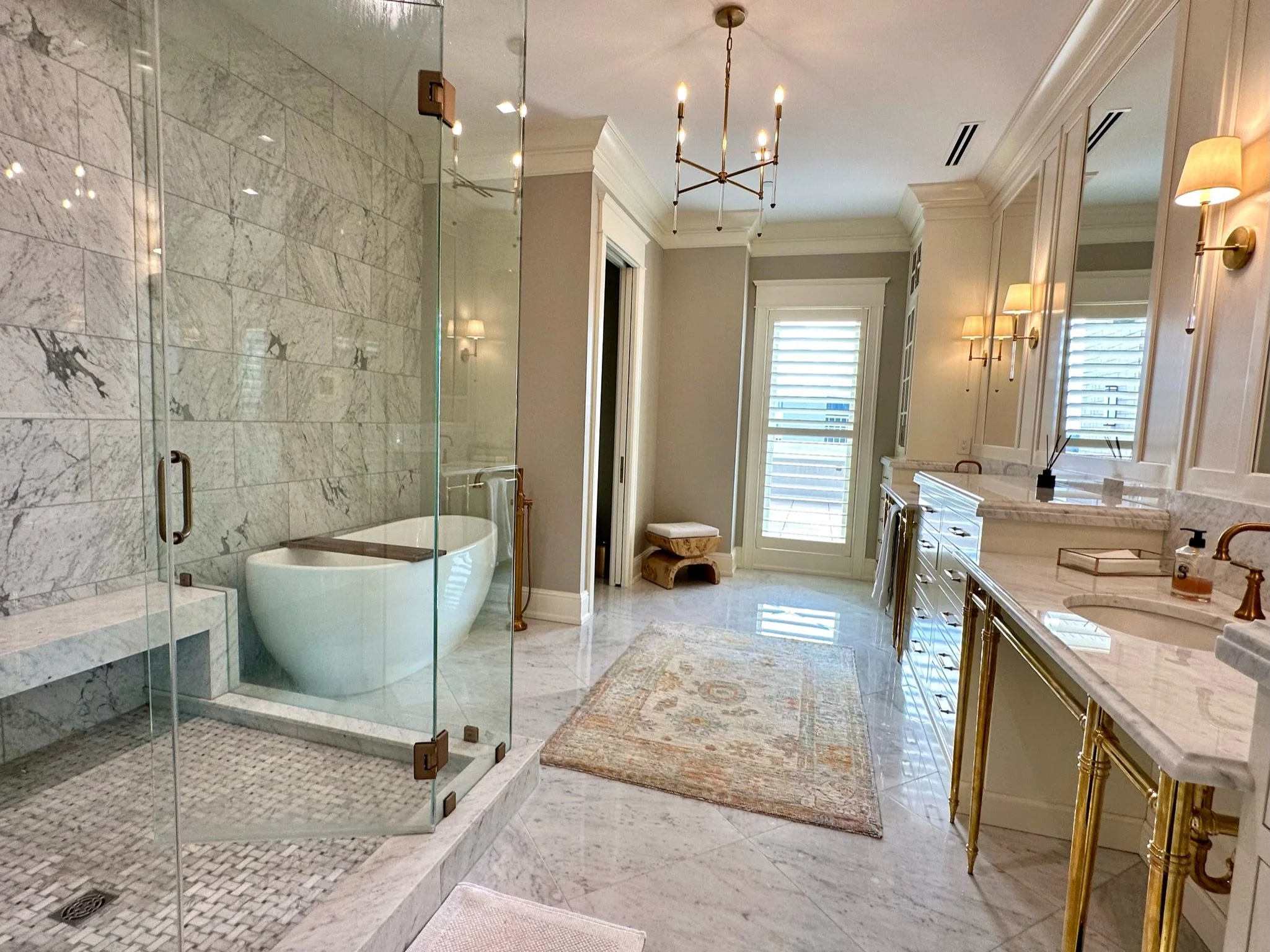 Luxurious bathroom featuring a glass-enclosed shower with marble tiles, a freestanding bathtub, large windows with shutters, elegant wall-mounted lamps, and a marble countertop vanity with gold accents.