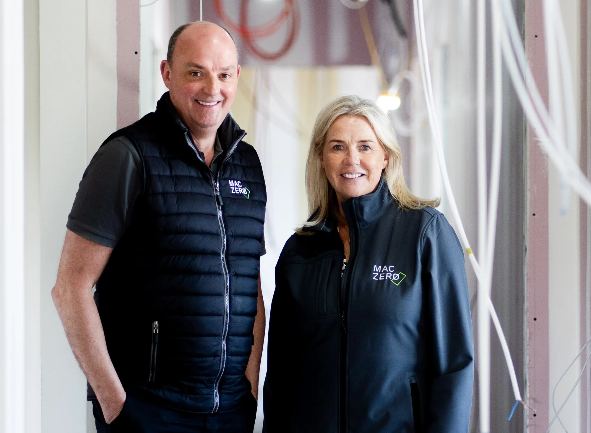 Two smiling people, a man and a woman, standing indoors next to a wall with electrical wires and pipes. Both are wearing black jackets with the 'MAC ZERO' logo.