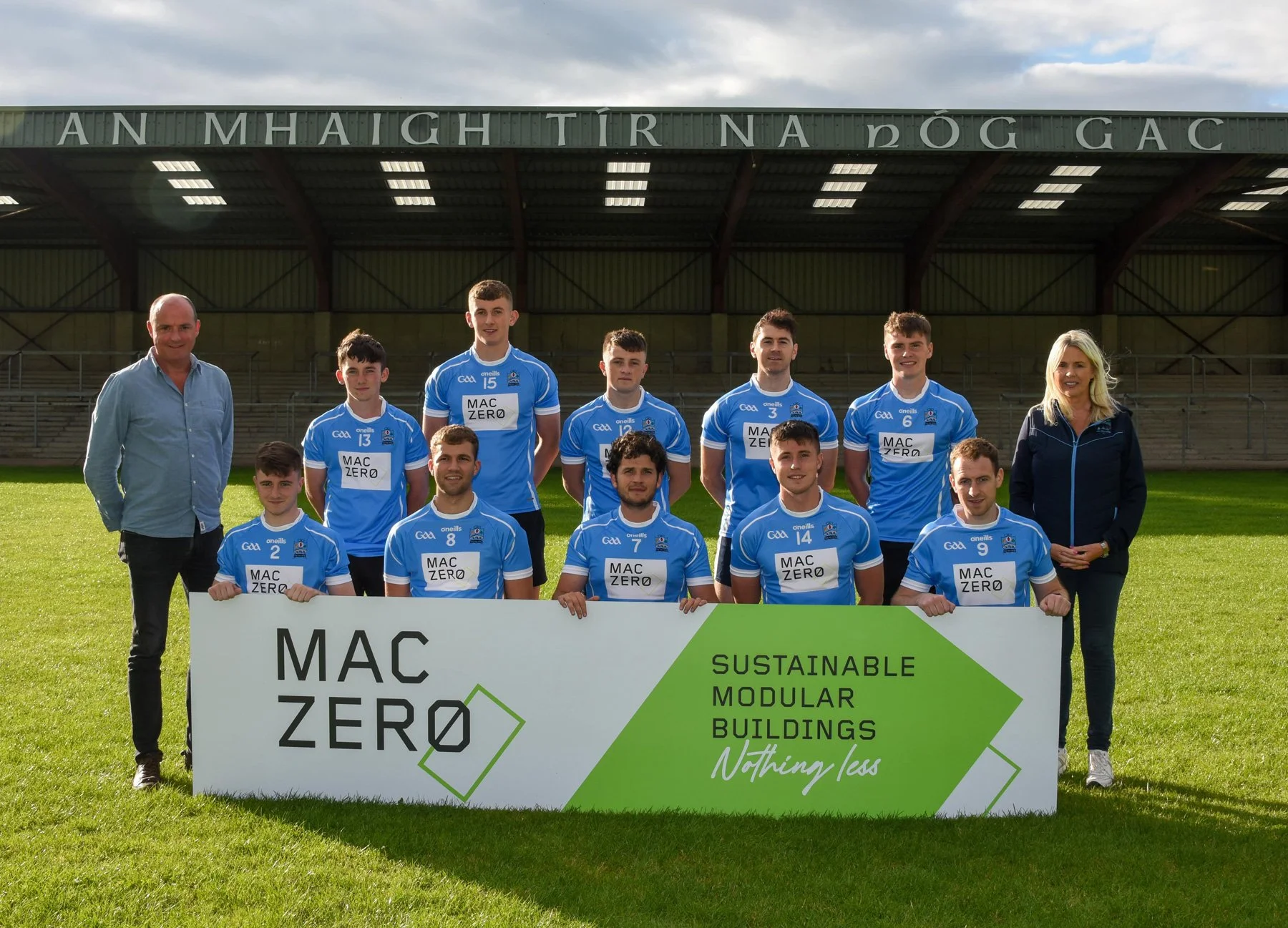 A group of young male athletes in blue sports jerseys with the logo GAA and sponsors, posing with two adults, one man and one woman, on a grass field under a stadium roof. They are holding a large sign that reads 'MAC ZERO' and 'SUSTAINABLE MODULAR BUILDINGS Nothing less'.