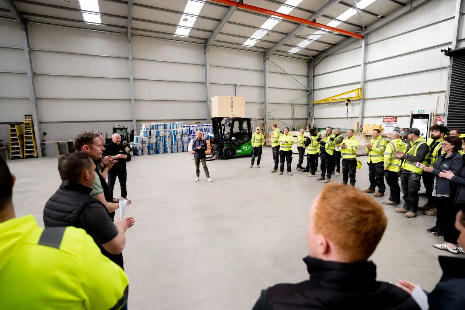 Group of workers in high-visibility vests inside a warehouse, standing in a semicircle around a woman speaking, with forklift and stacked boxes in the background.