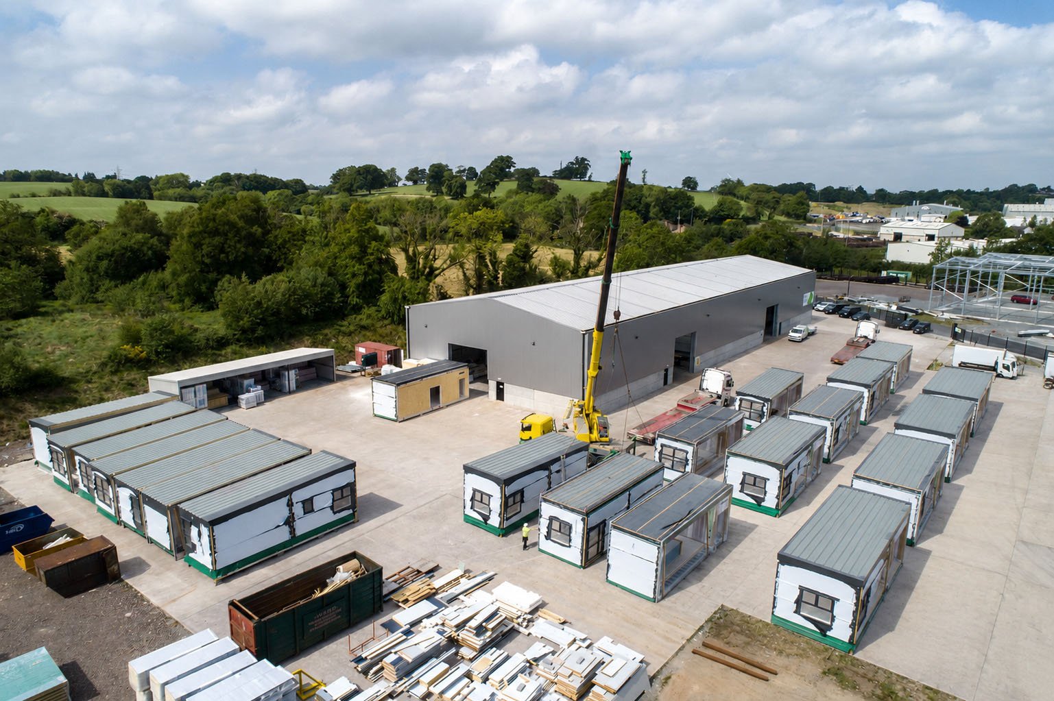 Aerial view of a construction site with multiple temporary structures, a large warehouse, parked trucks, and a crane lifting building materials, surrounded by green fields and some trees.