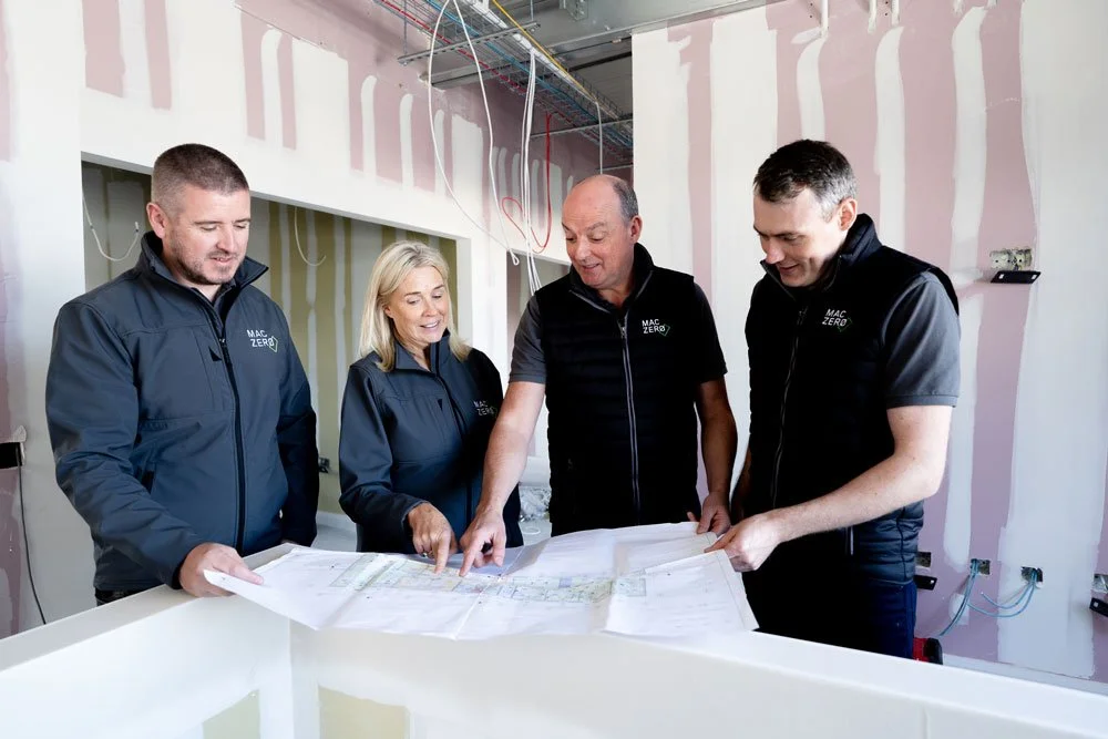Four people in black vests with 'MAC ZERO' logo examining blueprints inside a building under construction.