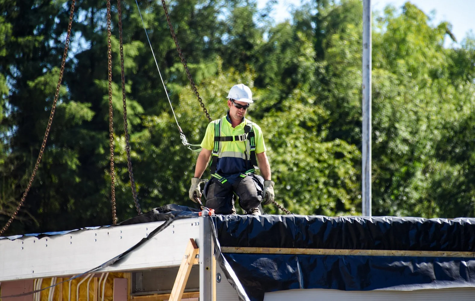 A construction worker wearing a white helmet, sunglasses, and a neon yellow safety vest is on a building site, working on the roof that is covered with black waterproofing material, with trees in the background.
