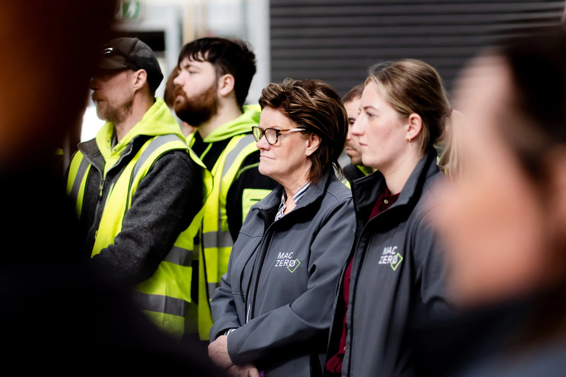 Group of people standing side by side, attending a presentation or meeting, some wearing jackets with 'MAC ZERO' logo, in an indoor setting.