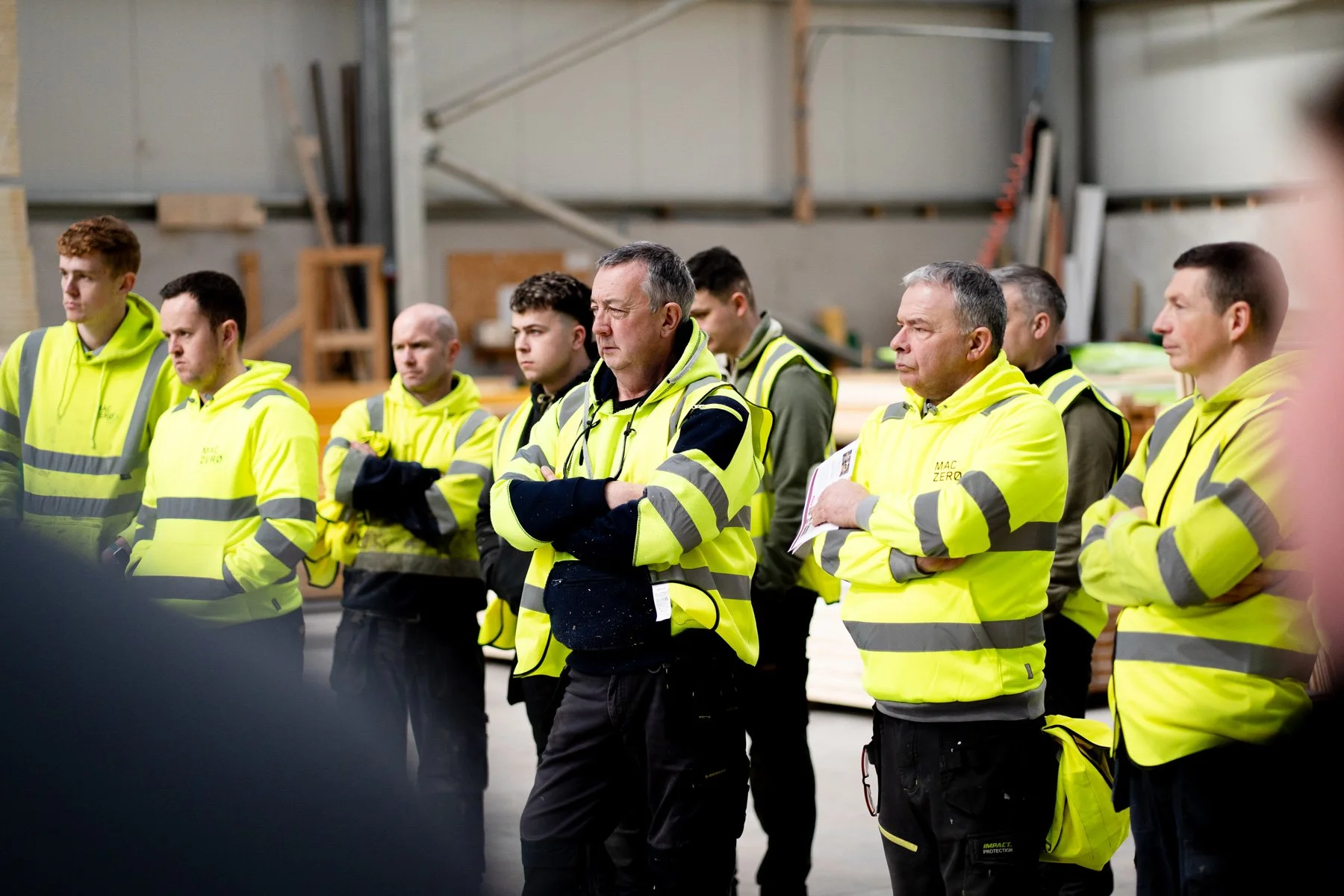 Group of industrial workers wearing yellow high-visibility jackets listening attentively in a workshop or warehouse.