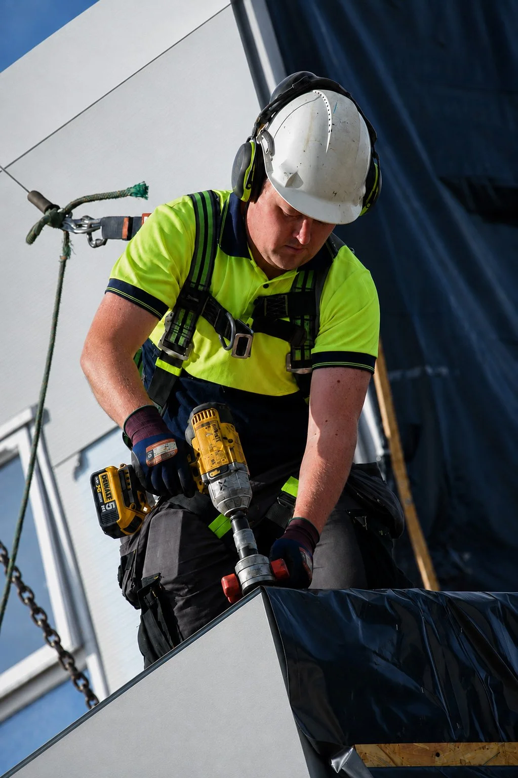 A construction worker wearing a white hard hat, high-visibility yellow shirt, and safety harness using a cordless drill on a building project outdoors.