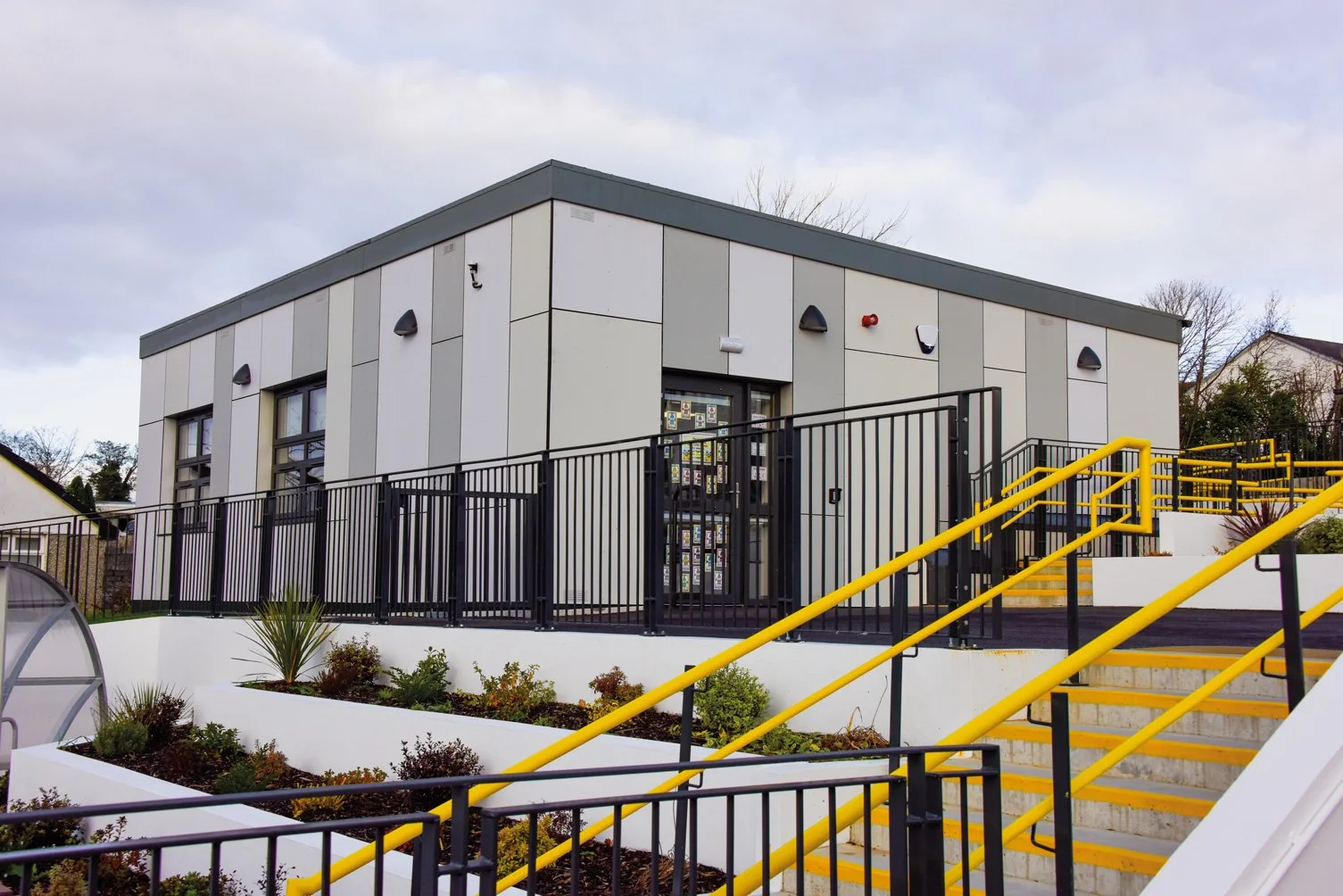 Two-story modern building with gray and white exterior panels, black railings, and yellow staircase handrails, surrounded by landscaped planters and outdoor stairs, under cloudy sky.