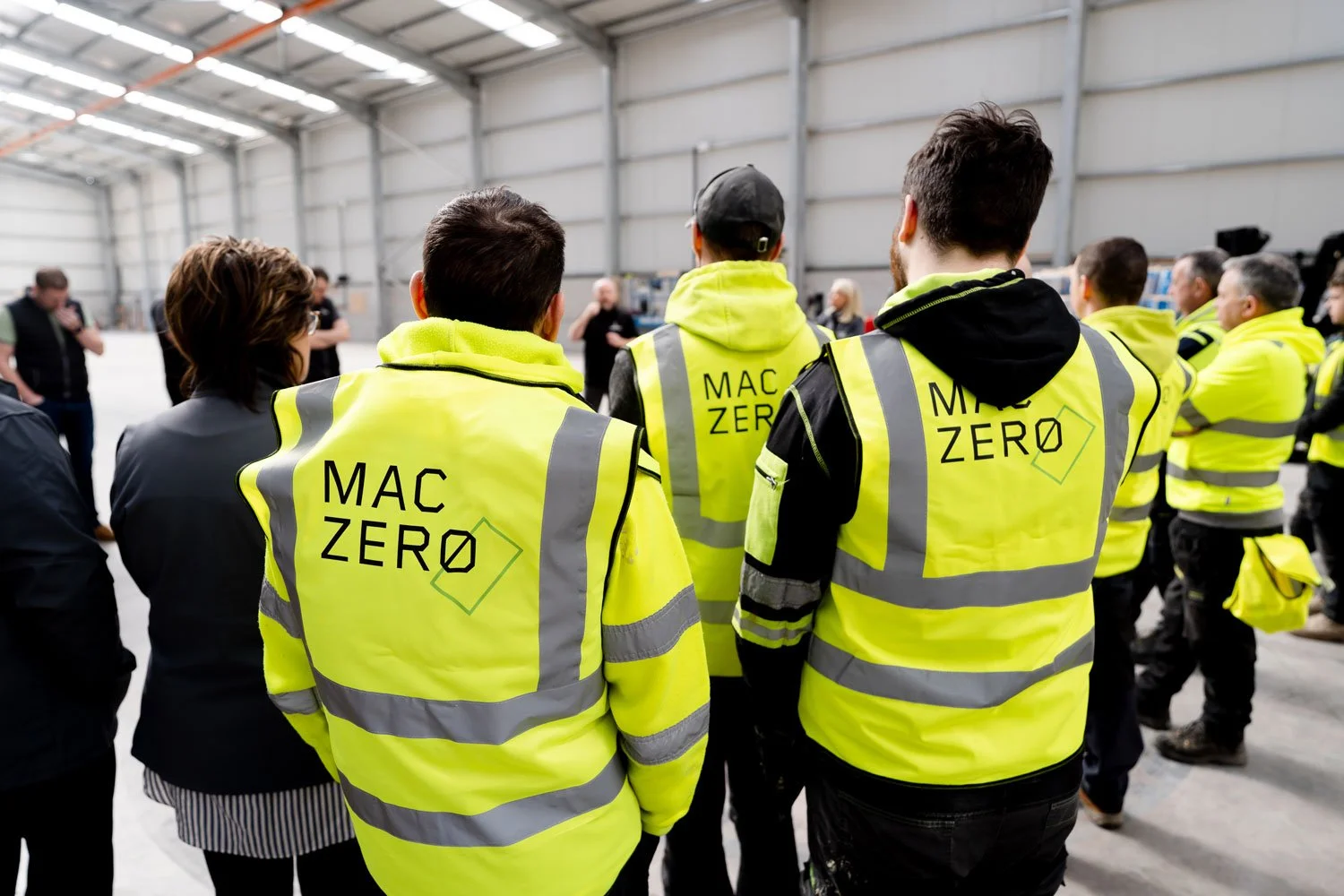 Group of people wearing yellow high-visibility vests with 'MAC ZERO' logo, gathered inside a large industrial building or warehouse, possibly attending a seminar or briefing.