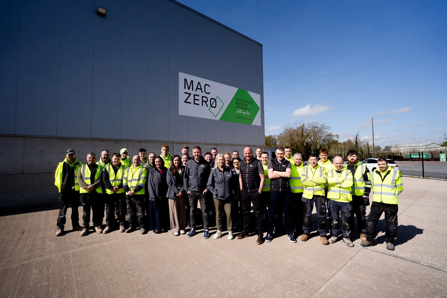 Group of people in yellow safety vests standing outside near a large gray building with a sign that reads 'MAC ZERO. Sustainable Modular Buildings.' The group is diverse in gender and age, and everyone is smiling.