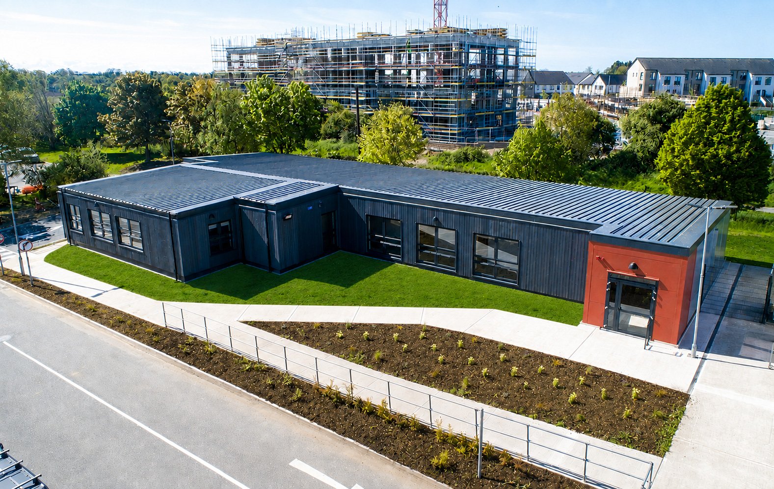 A modern, single-story building with a corrugated metal roof, black exterior walls, and a red accent wall at the entrance. It is surrounded by a landscaped lawn and walking paths, with an under-construction high-rise building and residential neighborhood in the background.