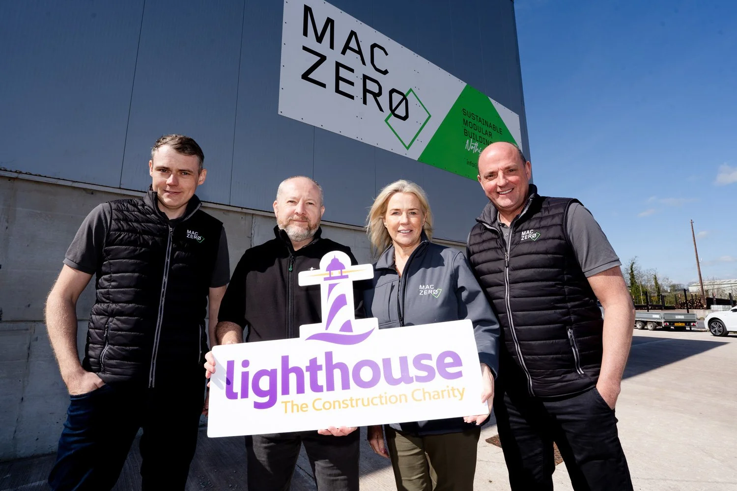 Four people standing outdoors in front of a building with a large sign that reads "MAC ZERO" and a smaller sign that says "Sustainable Modular Building". They are holding a sign that says "lighthouse The Construction Charity" with a lighthouse graphic. All are smiling and wearing jackets with the "MAC ZERO" logo.