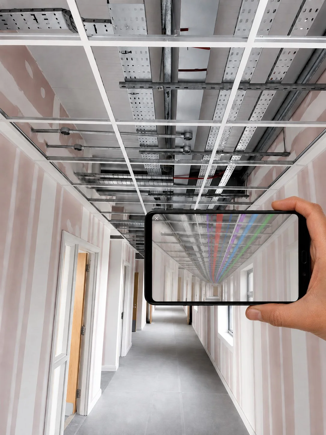 A person holding a smartphone captures a hallway with construction work on the ceiling, including metal framing and suspended lighting fixtures. The hallway has light-colored striped walls and multiple closed doors.