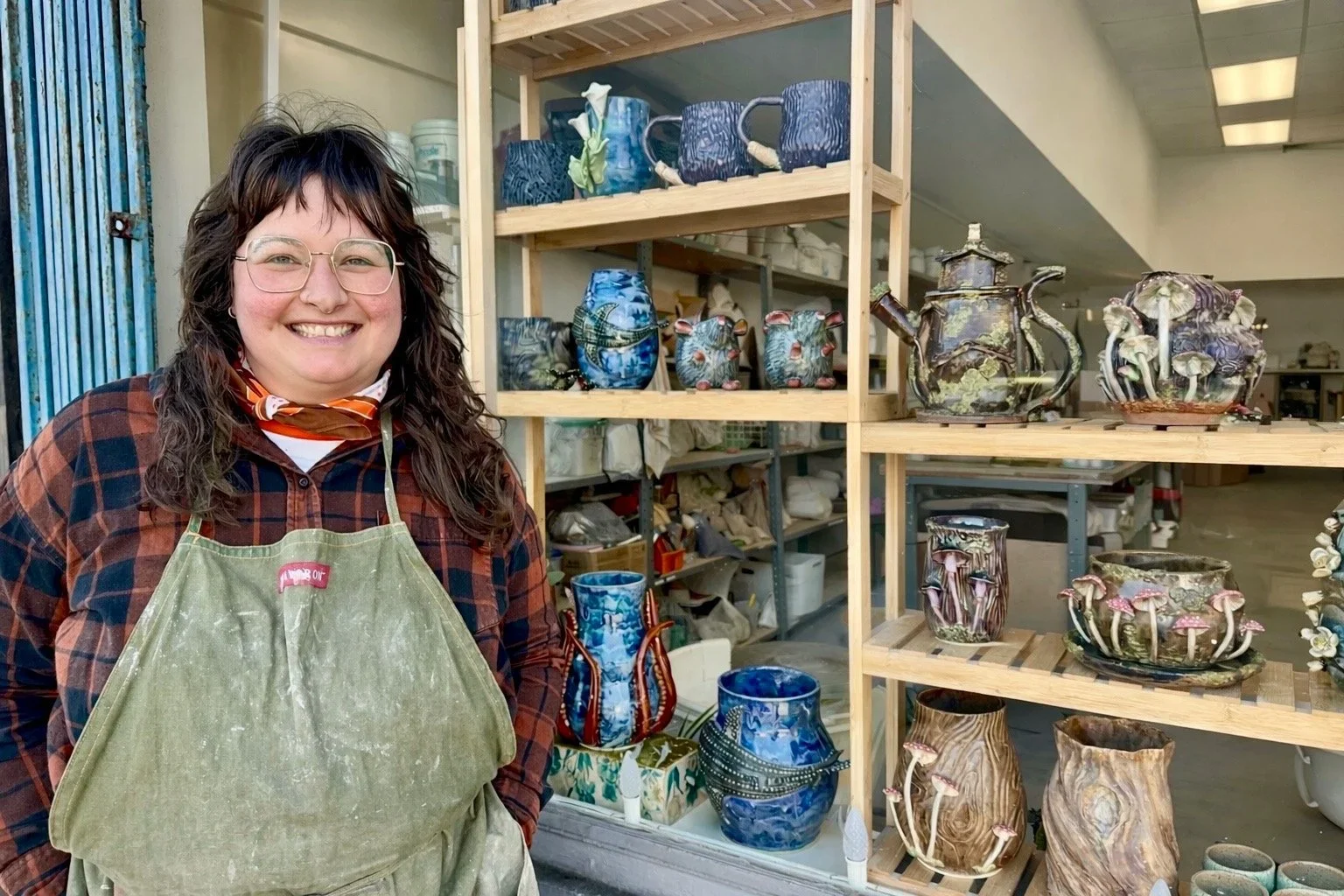 A smiling woman wearing glasses, a plaid shirt, and a green apron stands in front of shelves filled with colorful, handcrafted ceramic pots and teapots in a pottery studio.