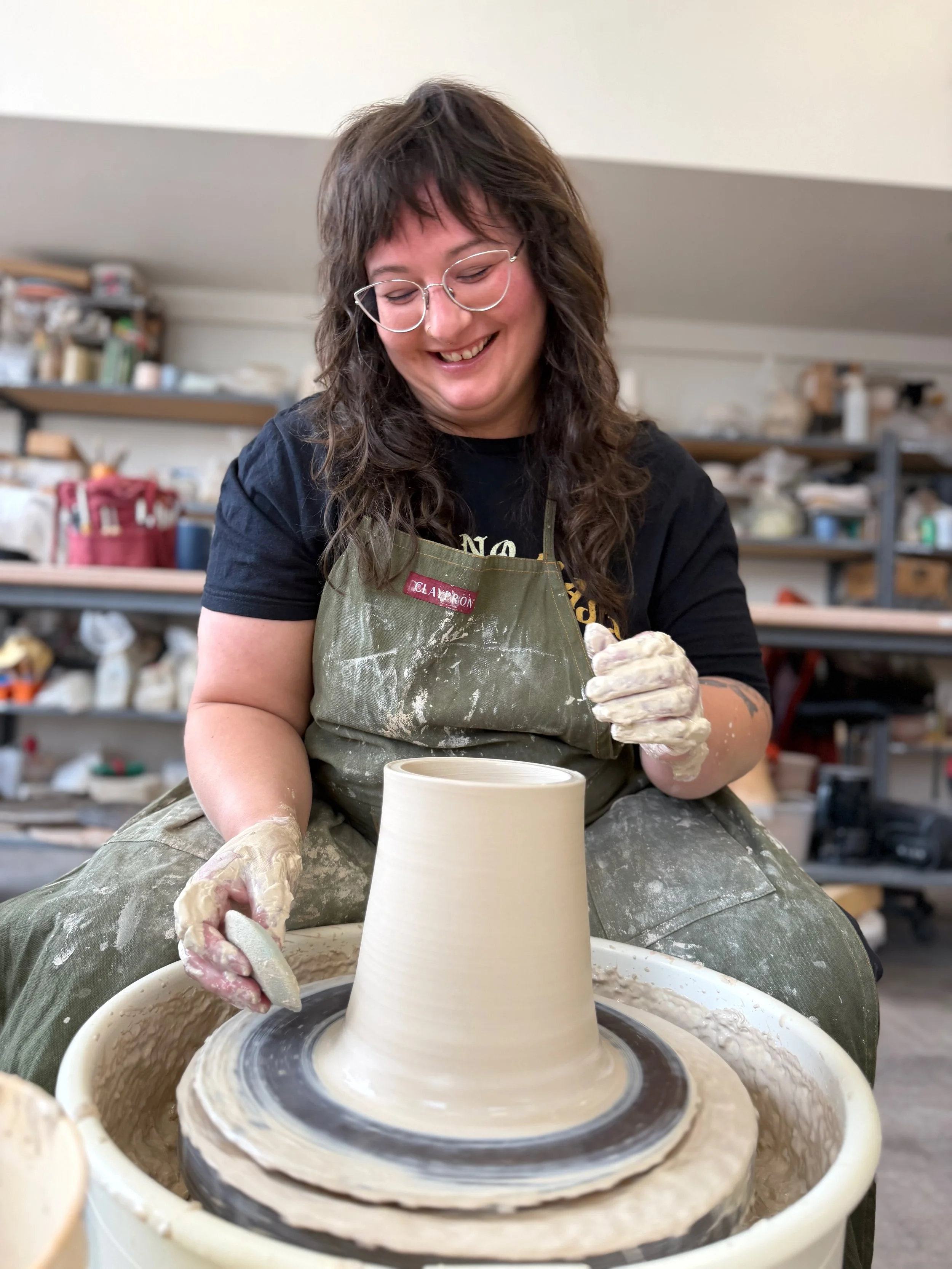 Woman with glasses and curly hair shaping a ceramic vase on a pottery wheel in a pottery studio.