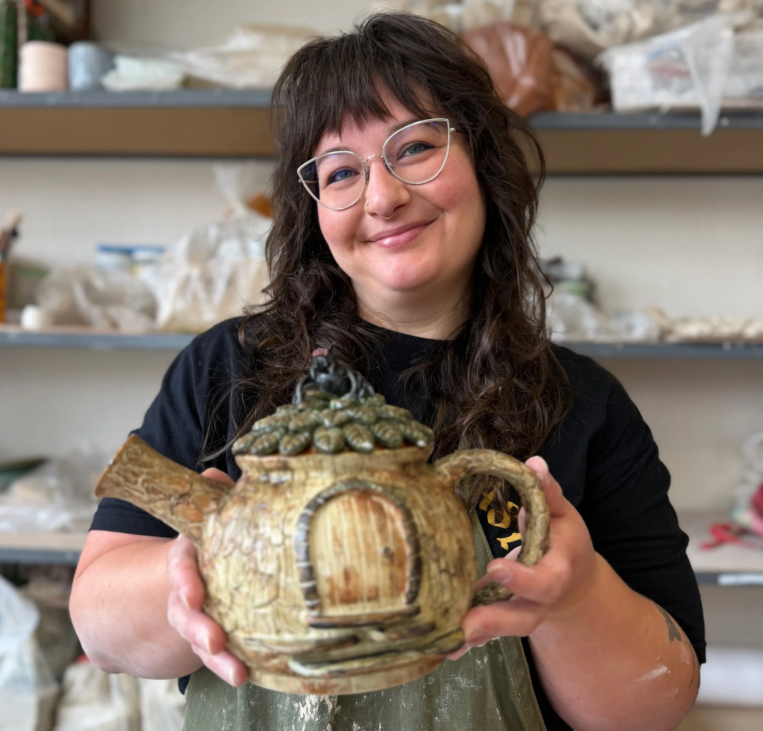 Smiling woman with glasses and tattoo holding a ceramic piece with small rounded details, in a workspace with shelves of ceramic materials in the background.