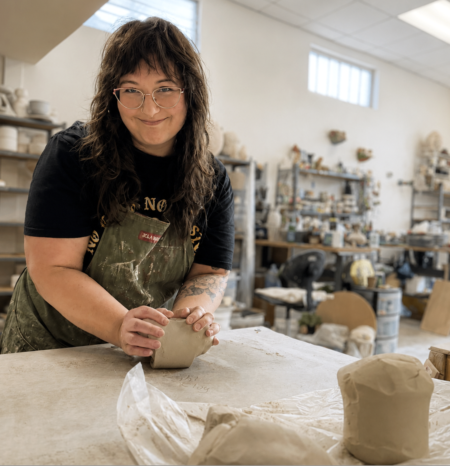A woman wearing glasses and a black t-shirt with a green apron is sculpting a clay piece in a ceramics studio. Shelves with pottery and art supplies are visible in the background.