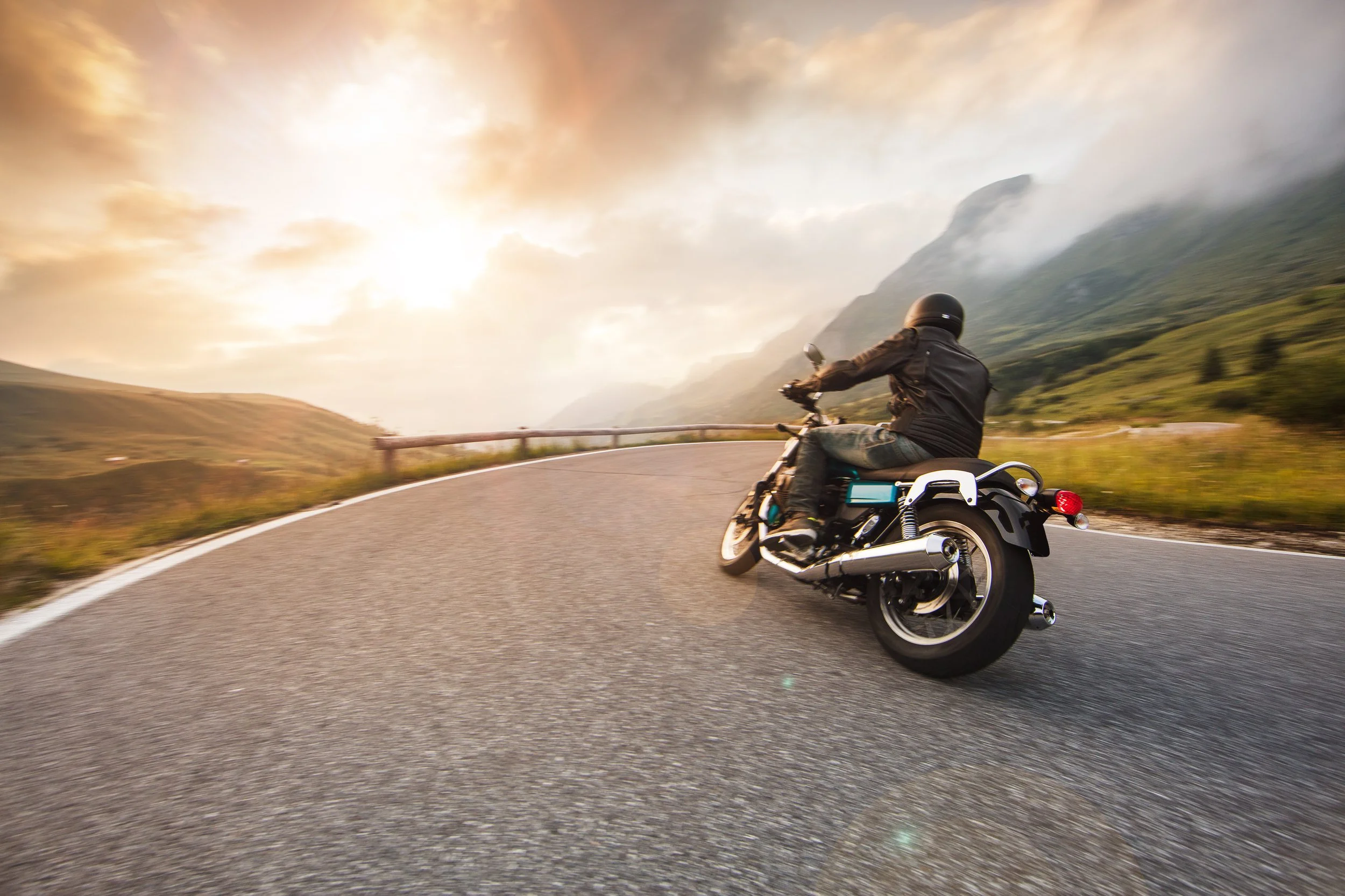 Motorcyclist riding on a curved mountain road during sunset or sunrise with misty mountains in the background.