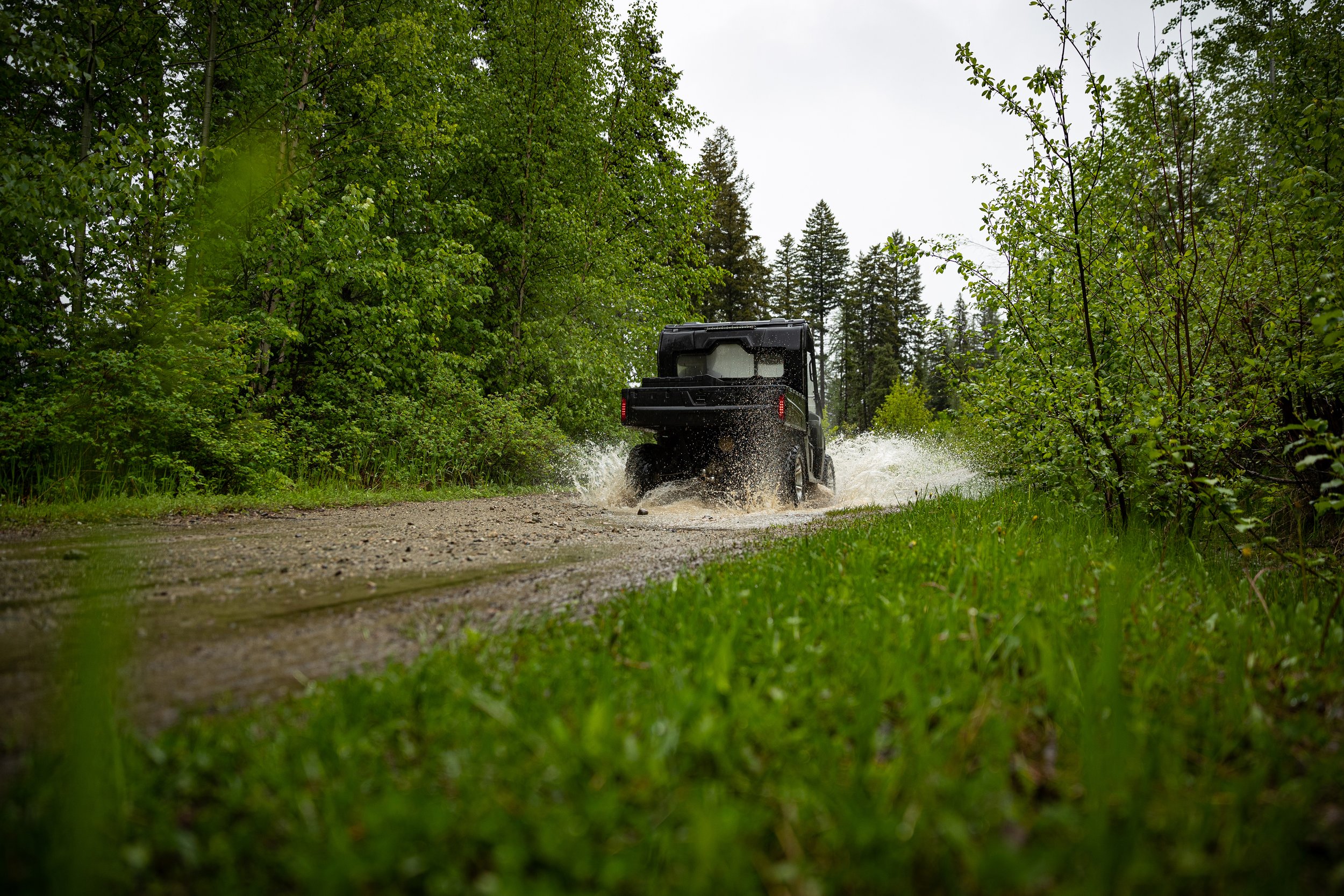 A black off-road vehicle driving through a muddy forest trail, splashing water on either side surrounded by green trees.