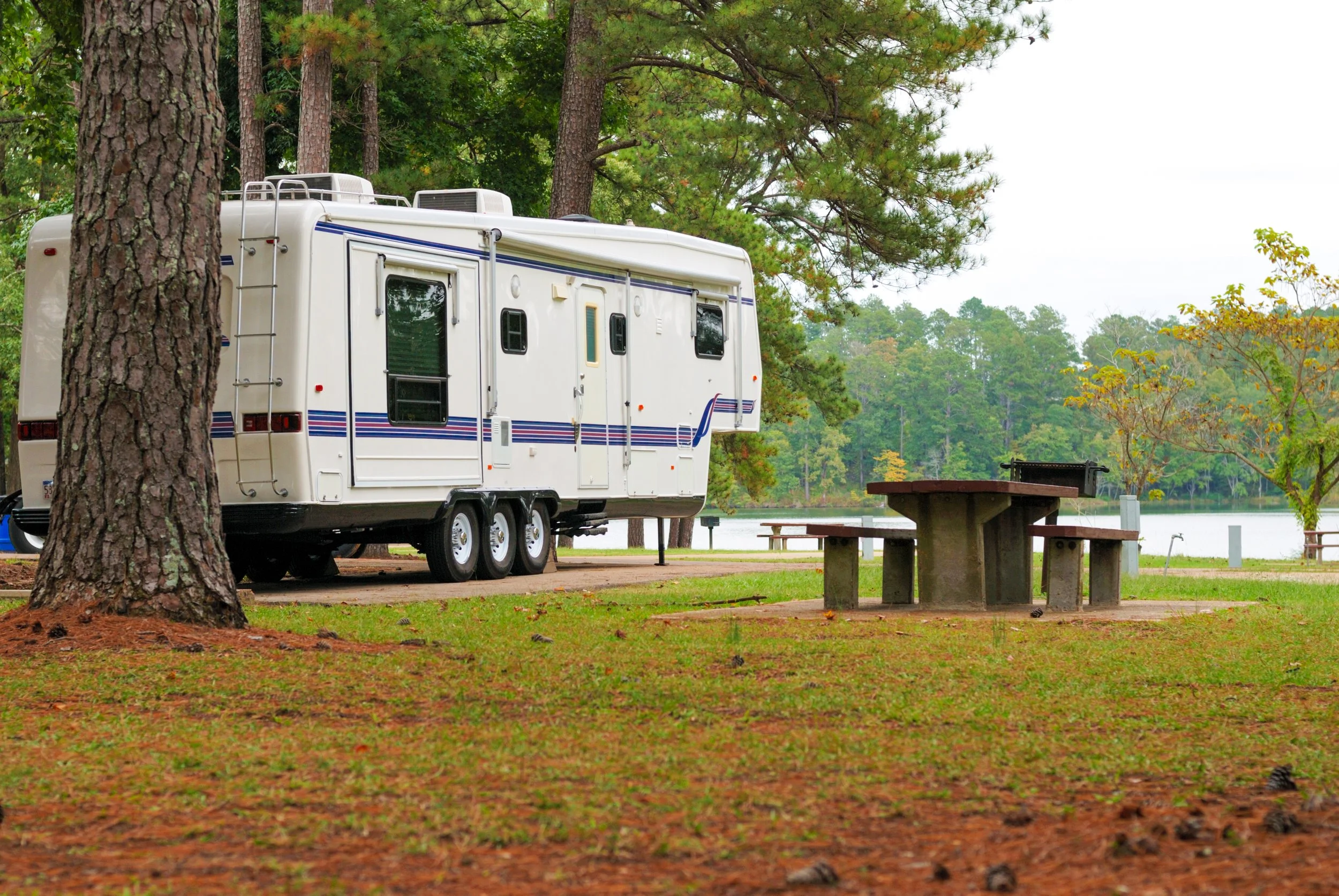 A white recreational vehicle parked on a dirt path in a wooded park near a lake, with picnic tables and trees around.