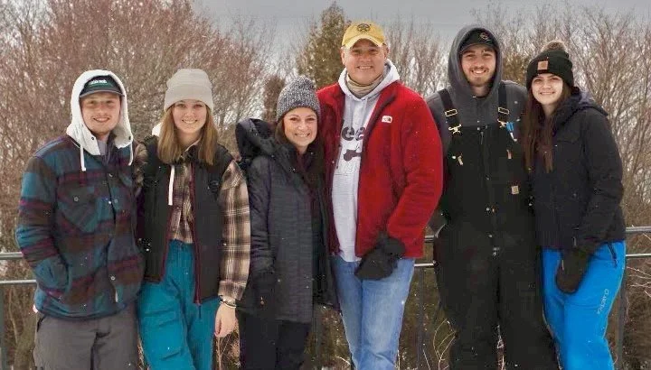 Group of six people standing outdoors in winter, dressed in warm clothing and hats, with trees and a fence in the background.