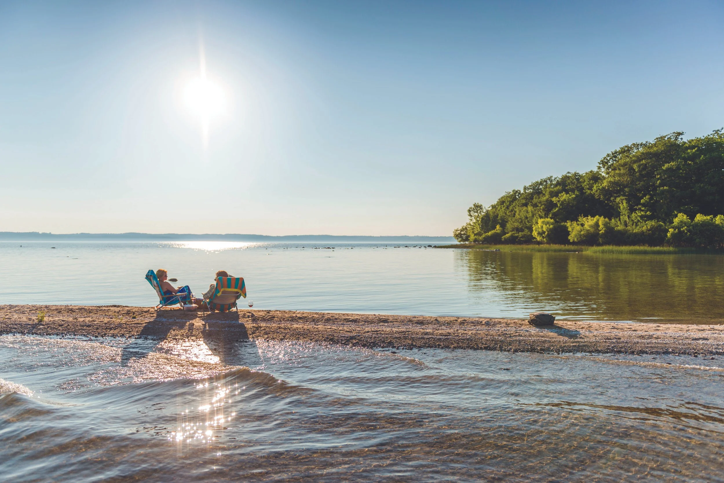 A peaceful lakeside scene on a sunny day with two people relaxing on chairs near the water, surrounded by trees and a clear sky.
