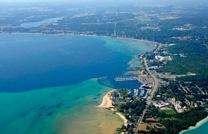 Aerial view of a lakeside town with a marina, sandy beach, and surrounding green areas, near a large body of water.