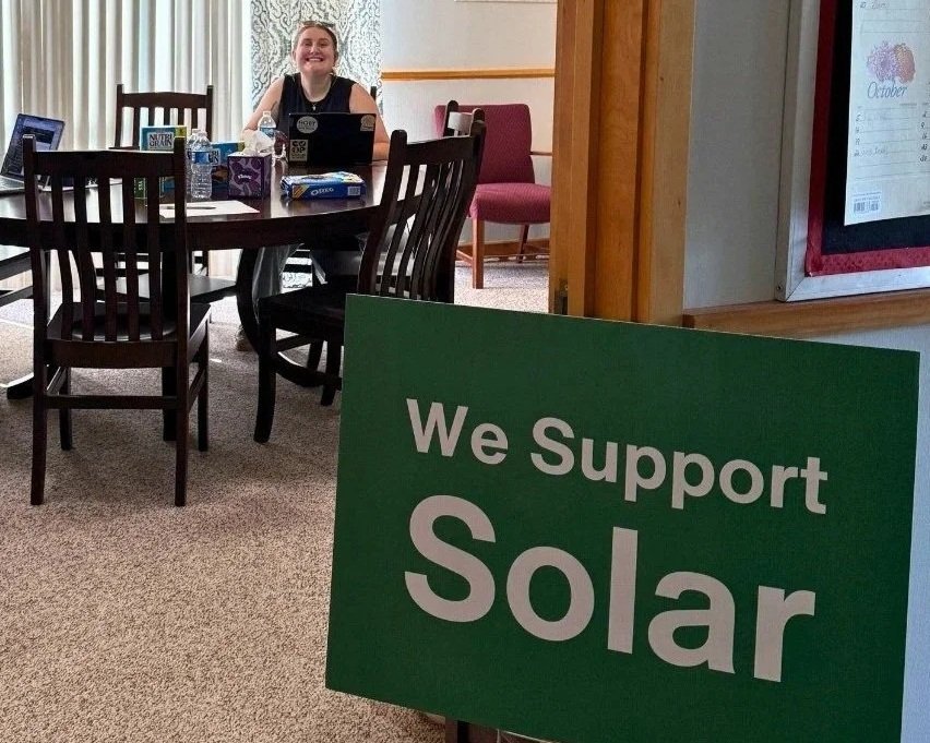 A woman sitting at a table in a room, smiling. The table has snacks, water bottles, and a laptop. In the foreground, there is a green sign that reads "We Support Solar."