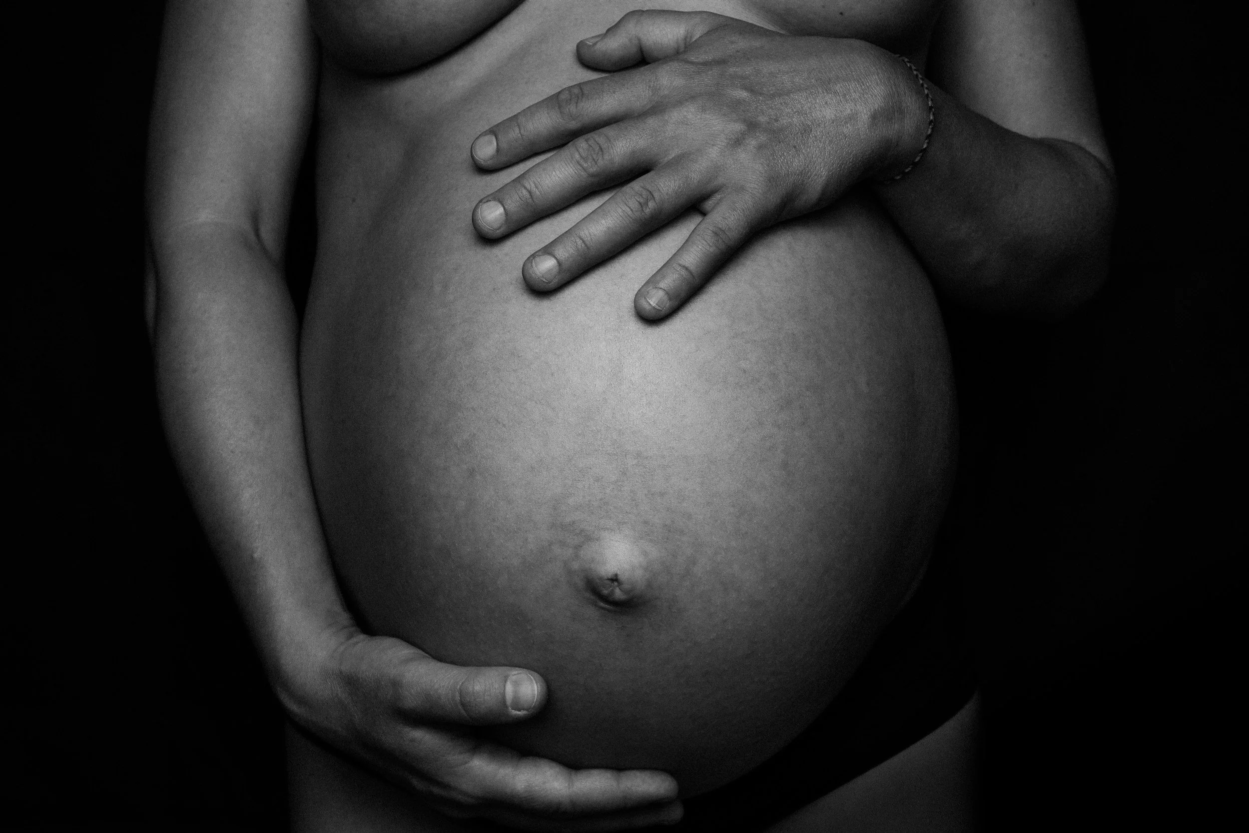 Black and white photograph of a person's hand gently resting on their pregnant belly, with the focus on the hand and belly area.