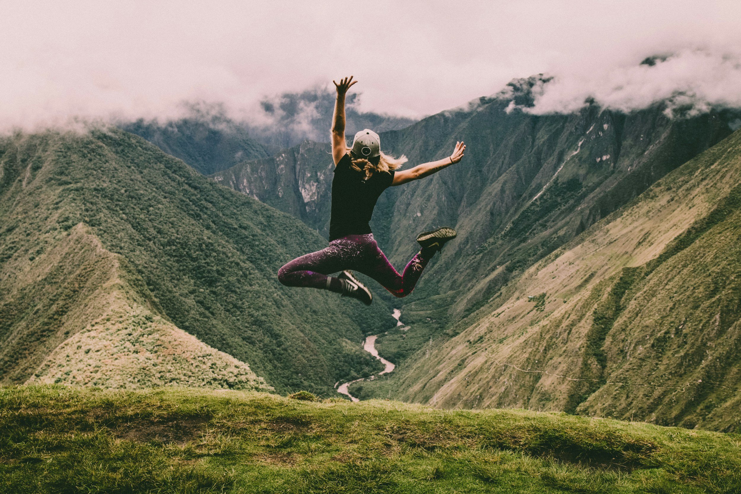 A person jumping in the air with arms raised, wearing a black shirt, patterned leggings, a baseball cap, and a harness, in a mountainous landscape with green slopes, a river, and cloudy skies.