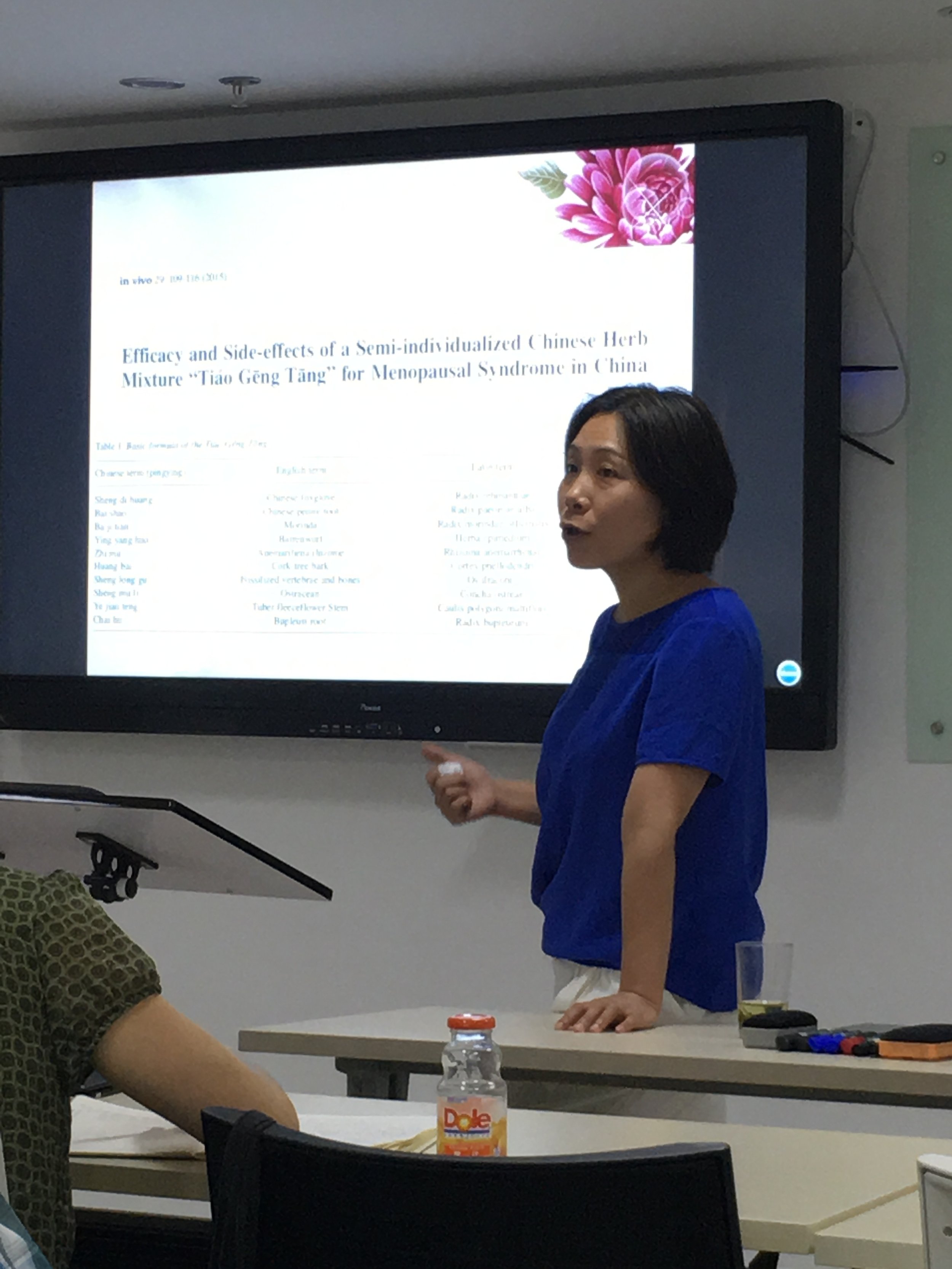 A woman in a blue shirt giving a presentation in a classroom. She is standing near a large screen displaying a research paper.