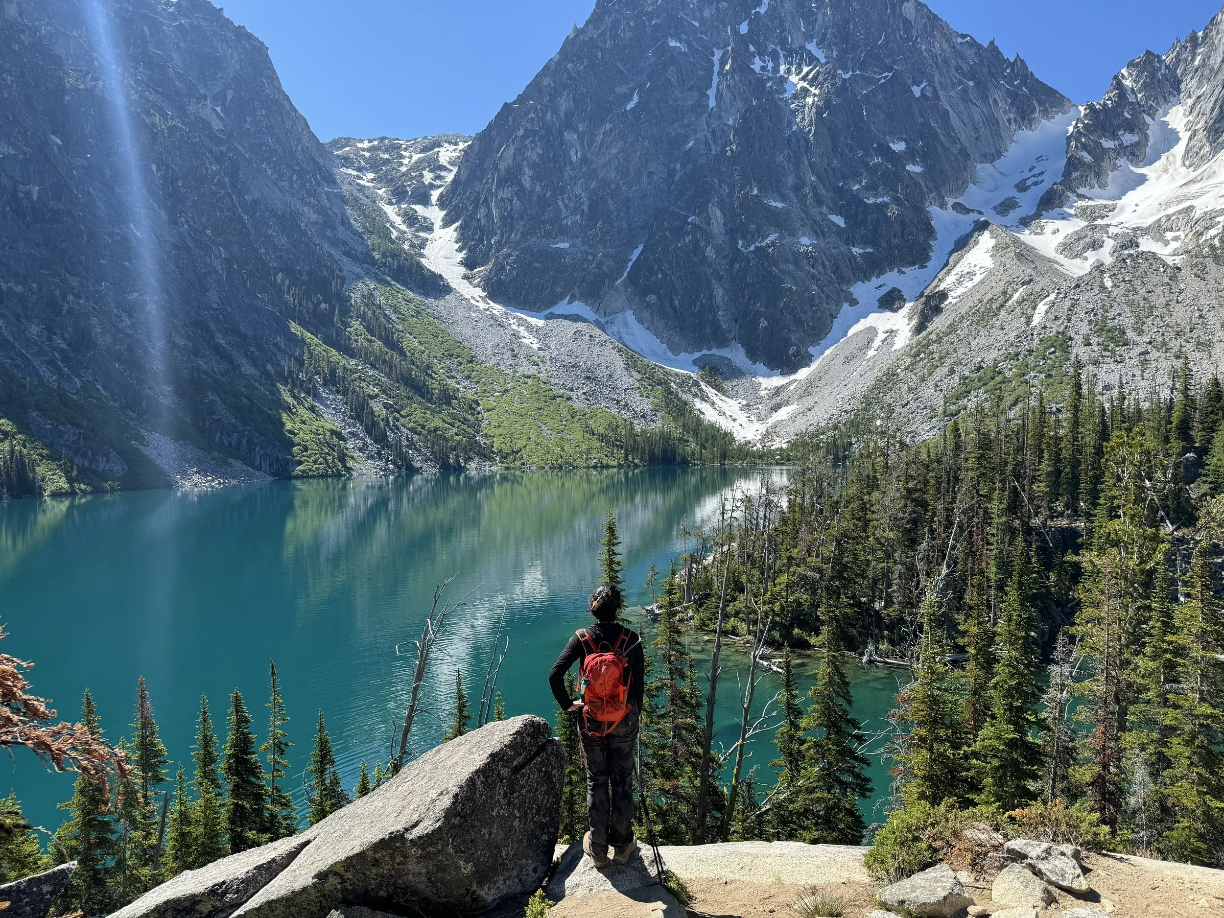 A hiker with a red backpack stands on a rocky trail, overlooking a turquoise mountain lake surrounded by dense pine trees and towering snow-capped mountains under a clear blue sky.