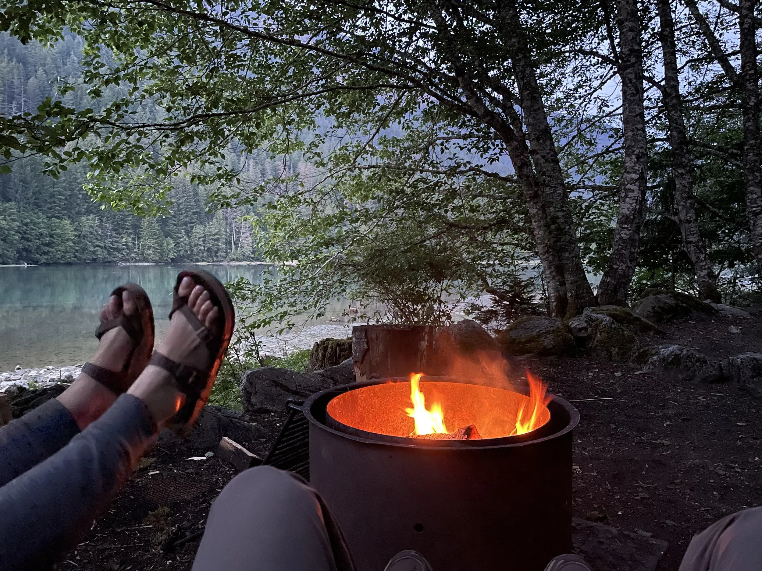 Person's feet in sandals resting near a campfire by a lake surrounded by trees and mountains.