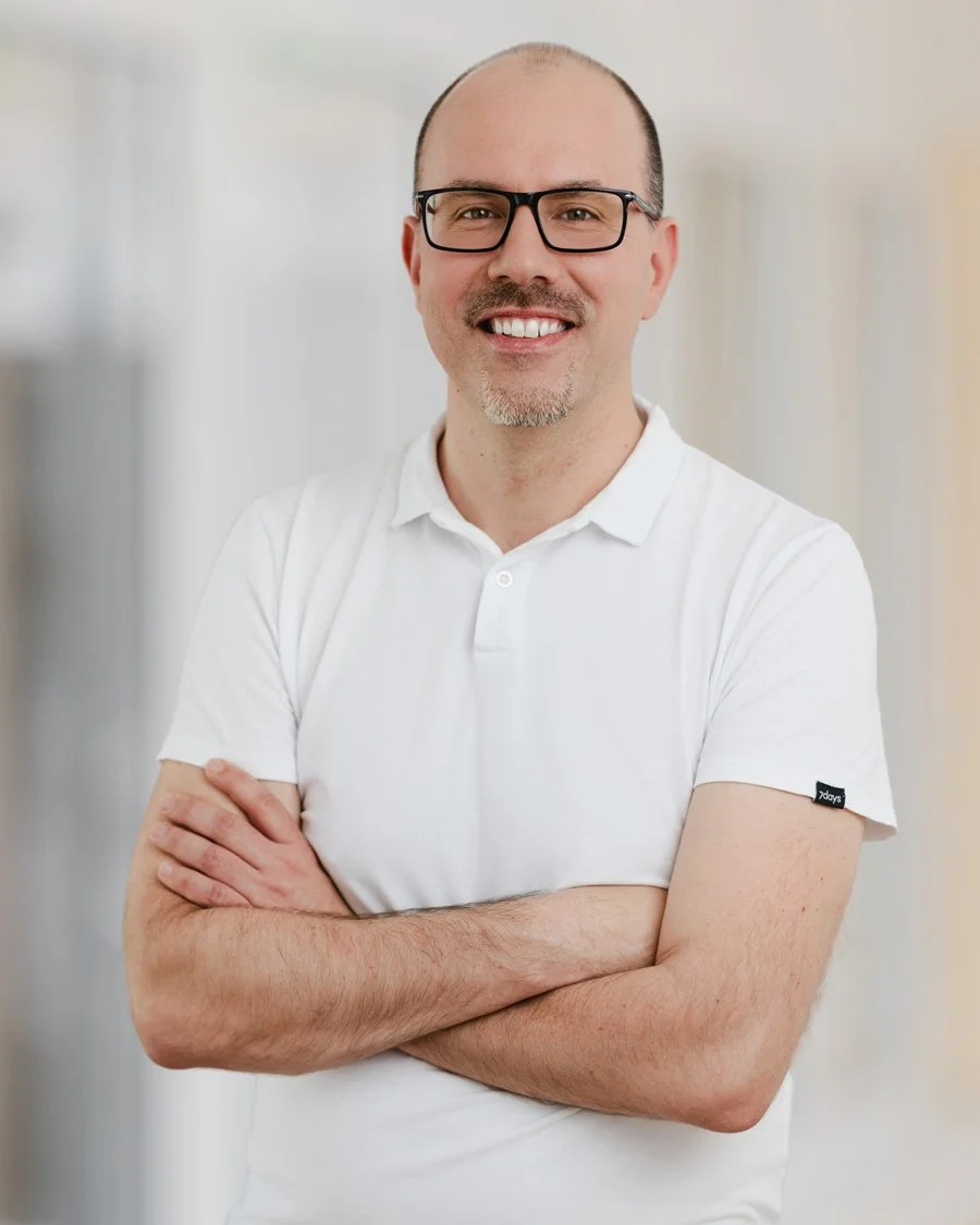 Lächelnder Mann mit Brille, weißem Poloshirt, kurz geschnittenem Haar und gepflegtem Bart, steht vor neutralem Hintergrund.
