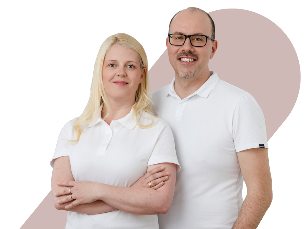 Stephanie Reichel und Dr. Andreas Reichel stehen nebeneinander, beide tragen weiße T-Shirts, lächeln und schauen in die Kamera, vor einem abstrakten Hintergrund.